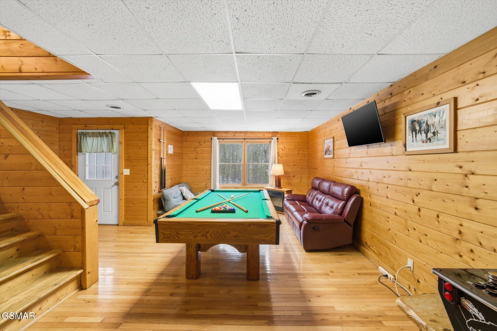 Wood-paneled game room with a pool table, leather reclining sofa, staircase, and pinball machine.