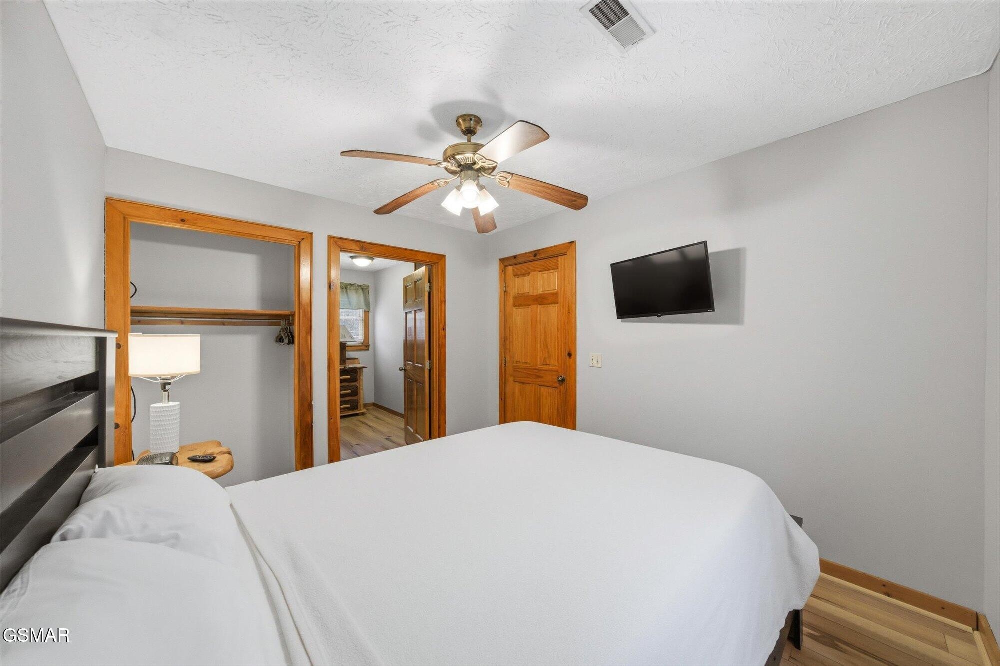 Bedroom with a white bed, wood-trimmed closet, wall-mounted television, and ceiling fan.