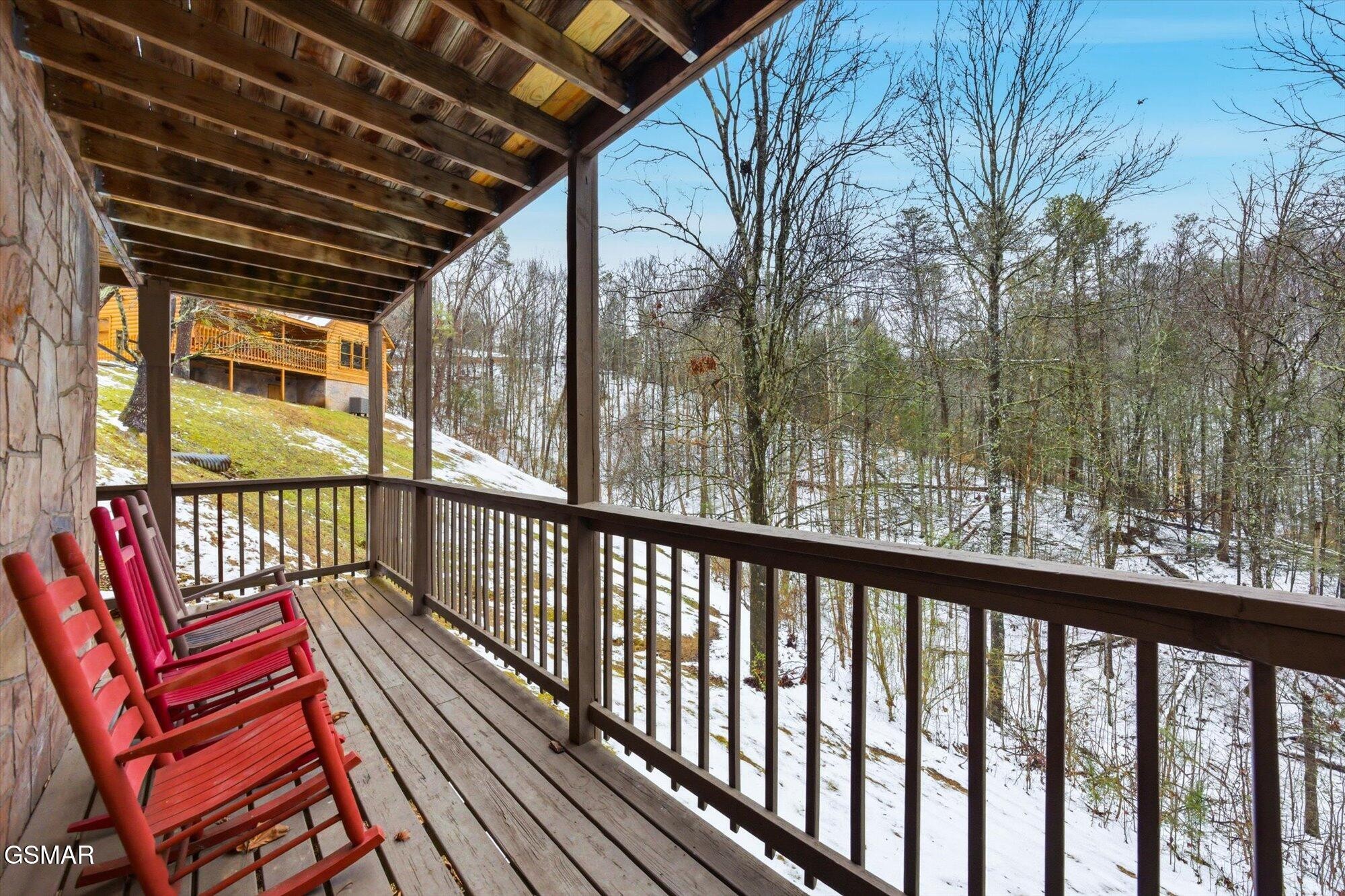 Covered wooden deck with red rocking chairs overlooking snowy woods and a neighboring cabin.