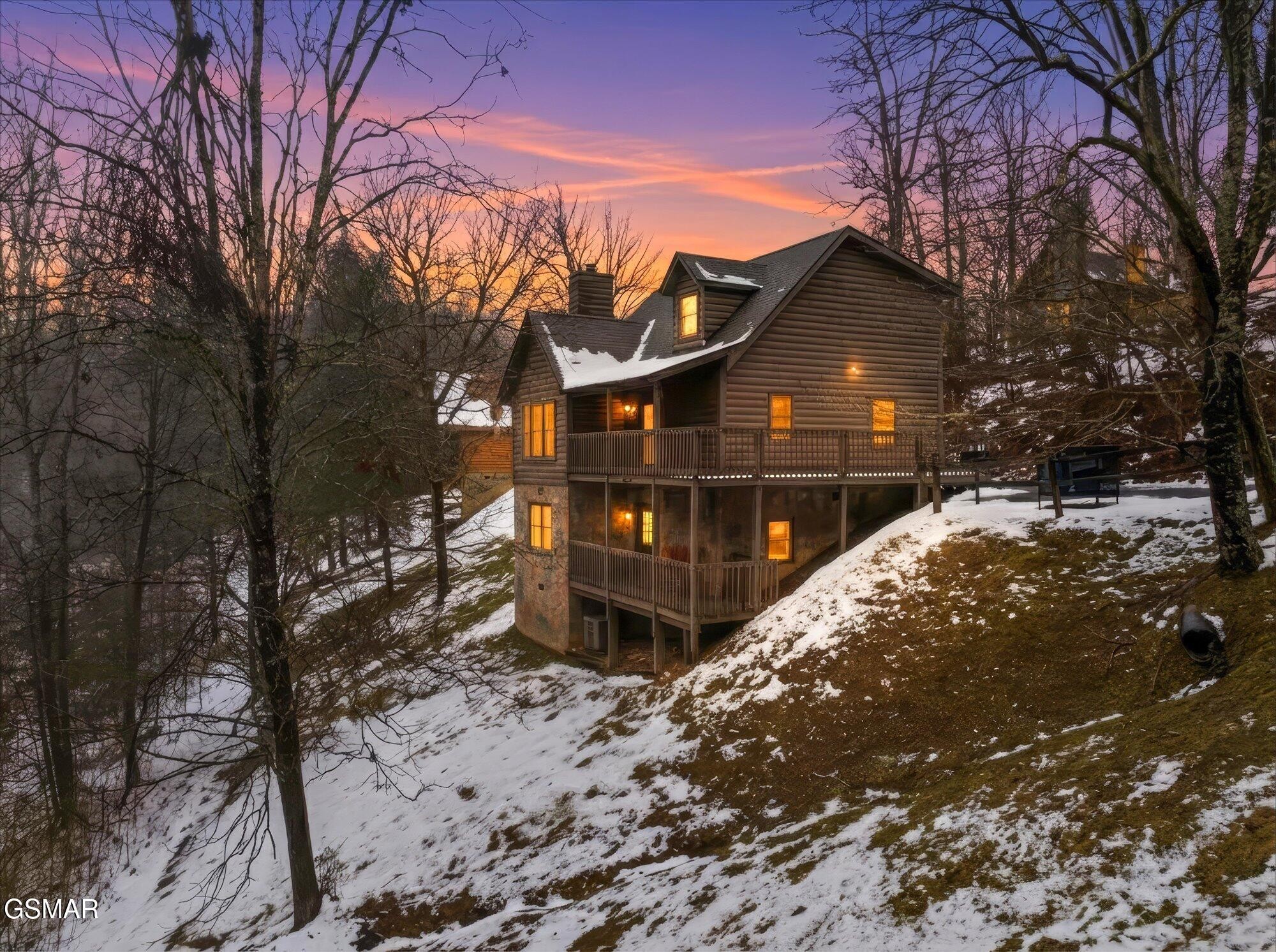 Multi-level log cabin with wraparound decks on a snowy wooded hillside at sunset.