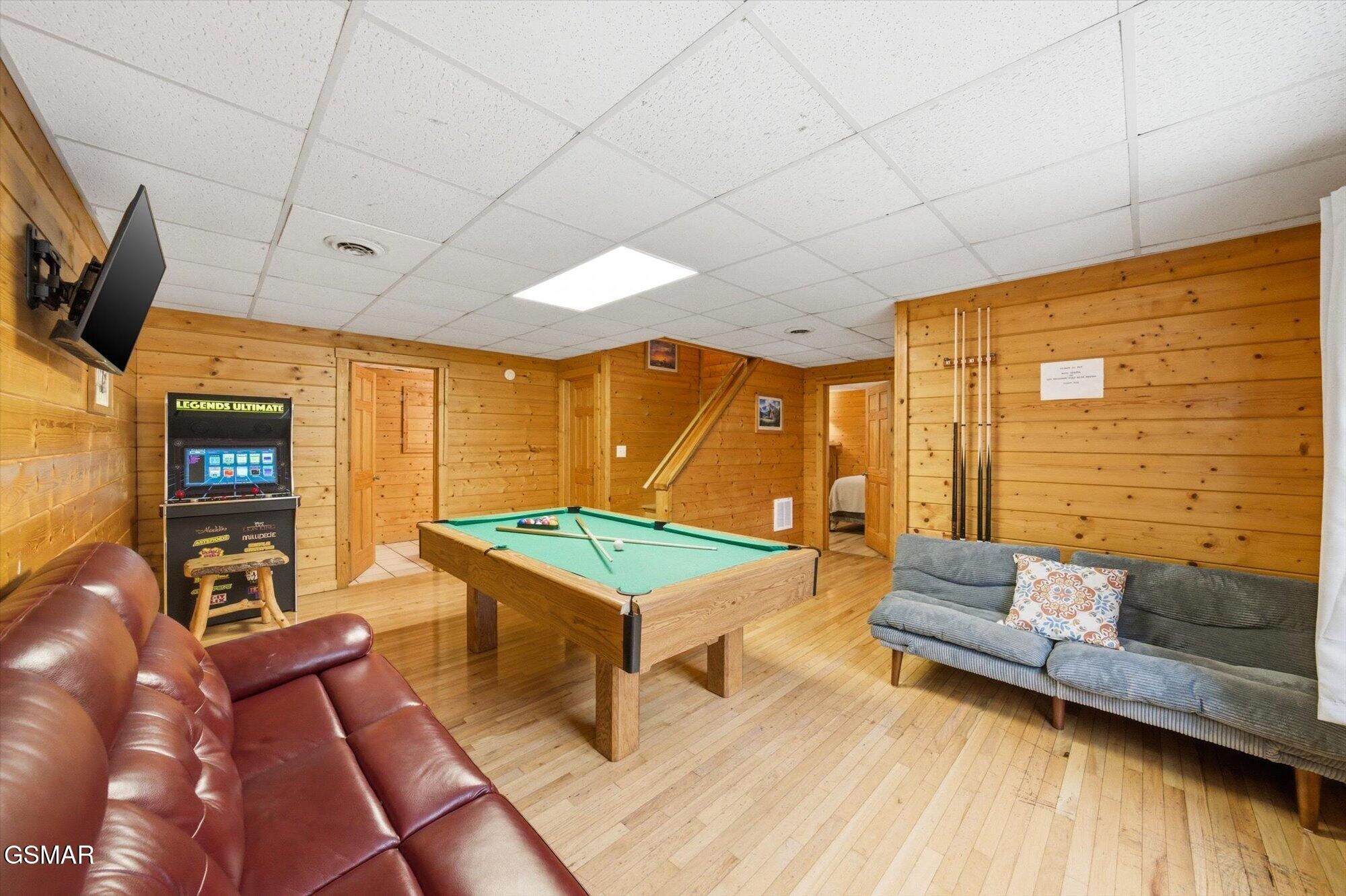 Wood-paneled game room with a pool table, arcade game, sofas, and stairs.