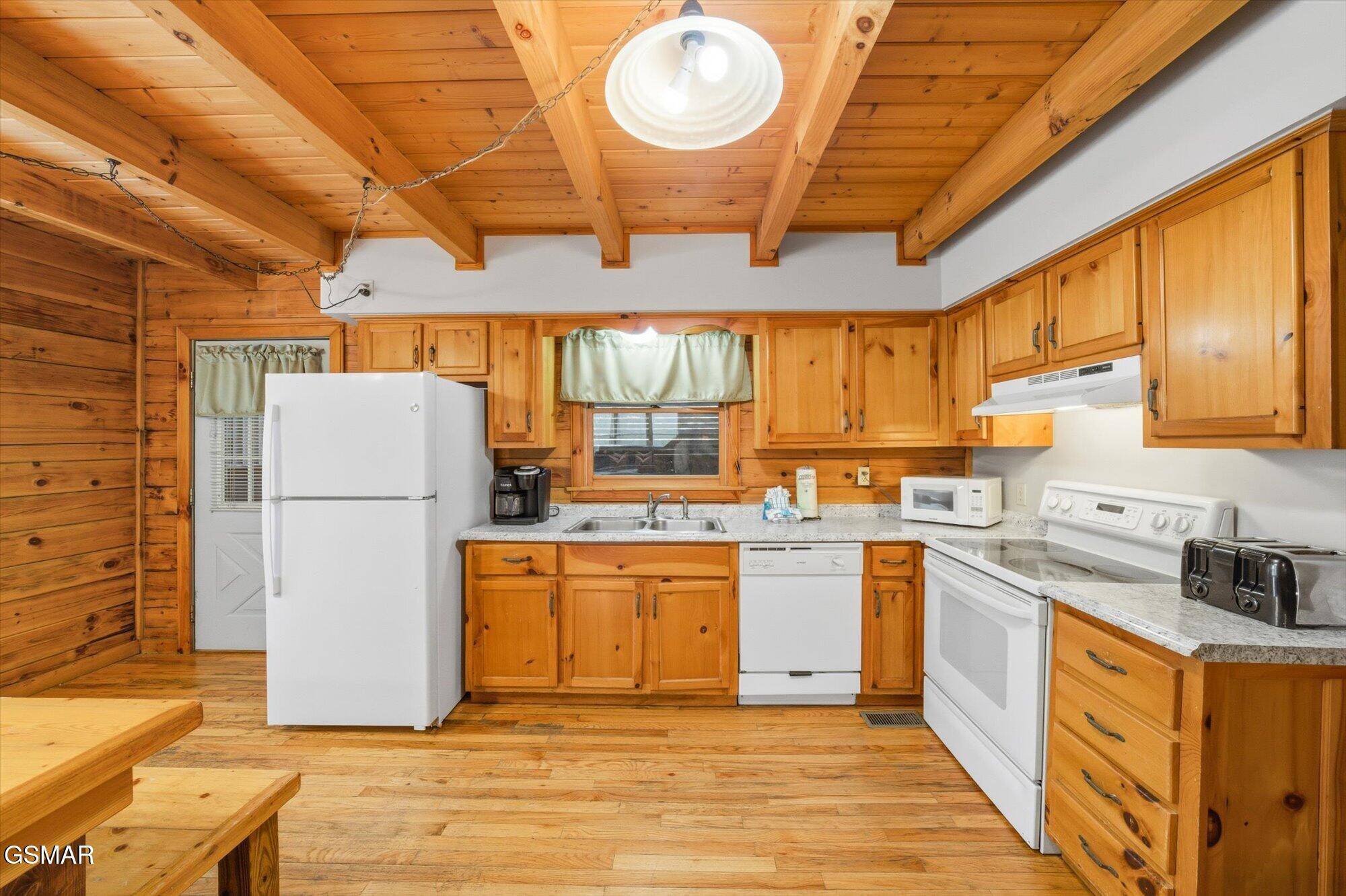 Wood-paneled kitchen with exposed ceiling beams, honey-colored cabinets, and white appliances.