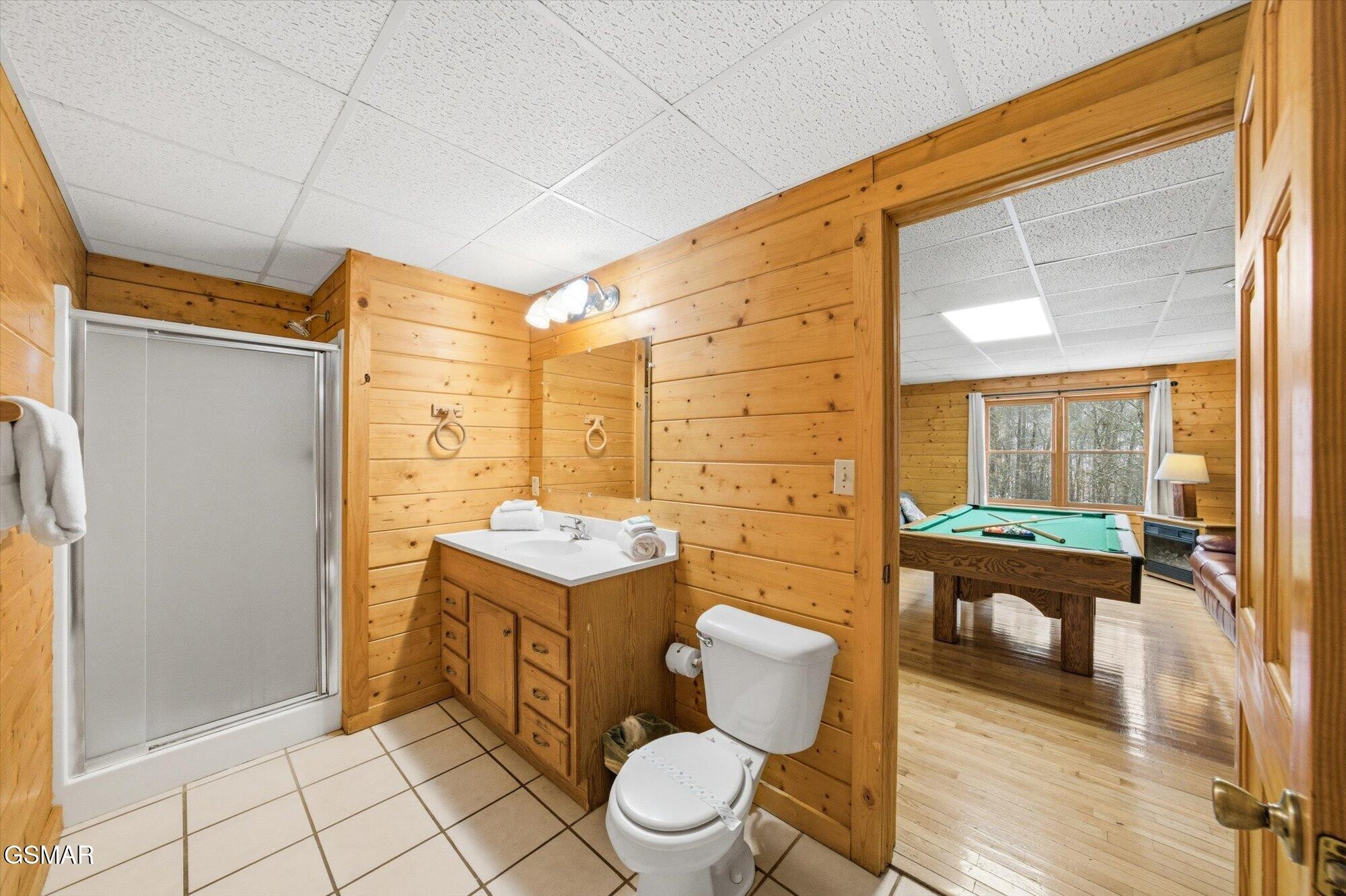 Wood-paneled bathroom with a tiled floor, shower, vanity, and a view into a game room.