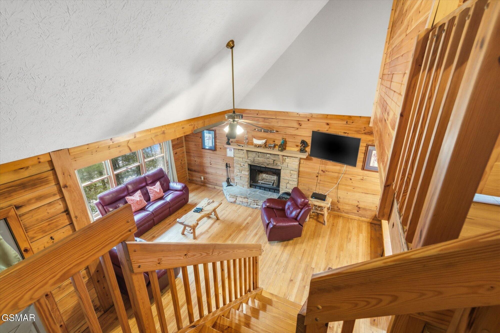 Cabin living room with wood-paneled walls, stone fireplace, and burgundy leather seating, viewed from the stairs.