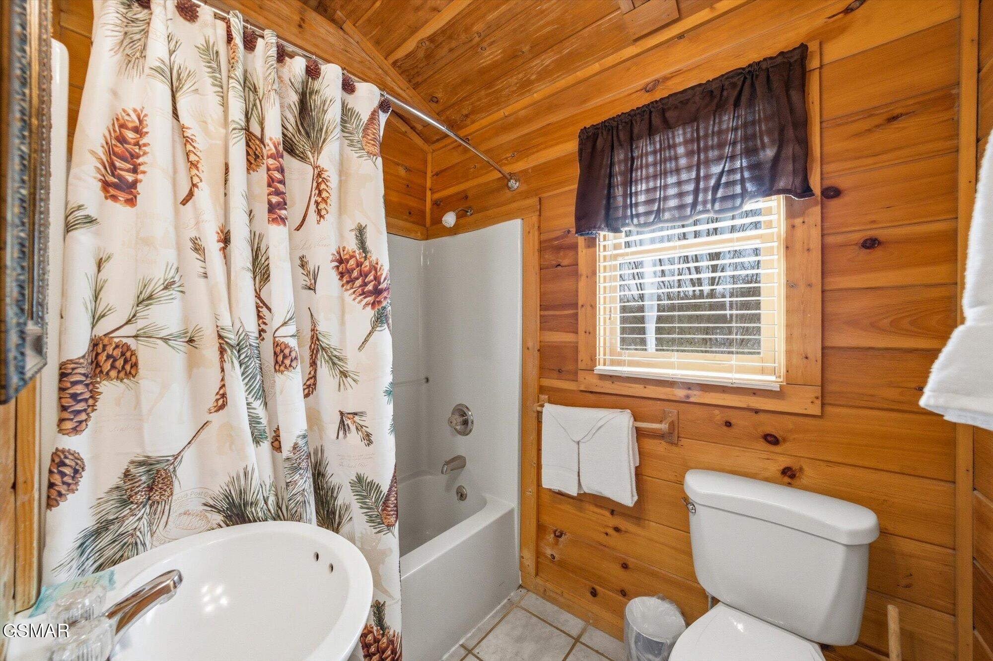 Rustic cabin bathroom with wood walls, a pine-cone shower curtain, bathtub, and toilet.