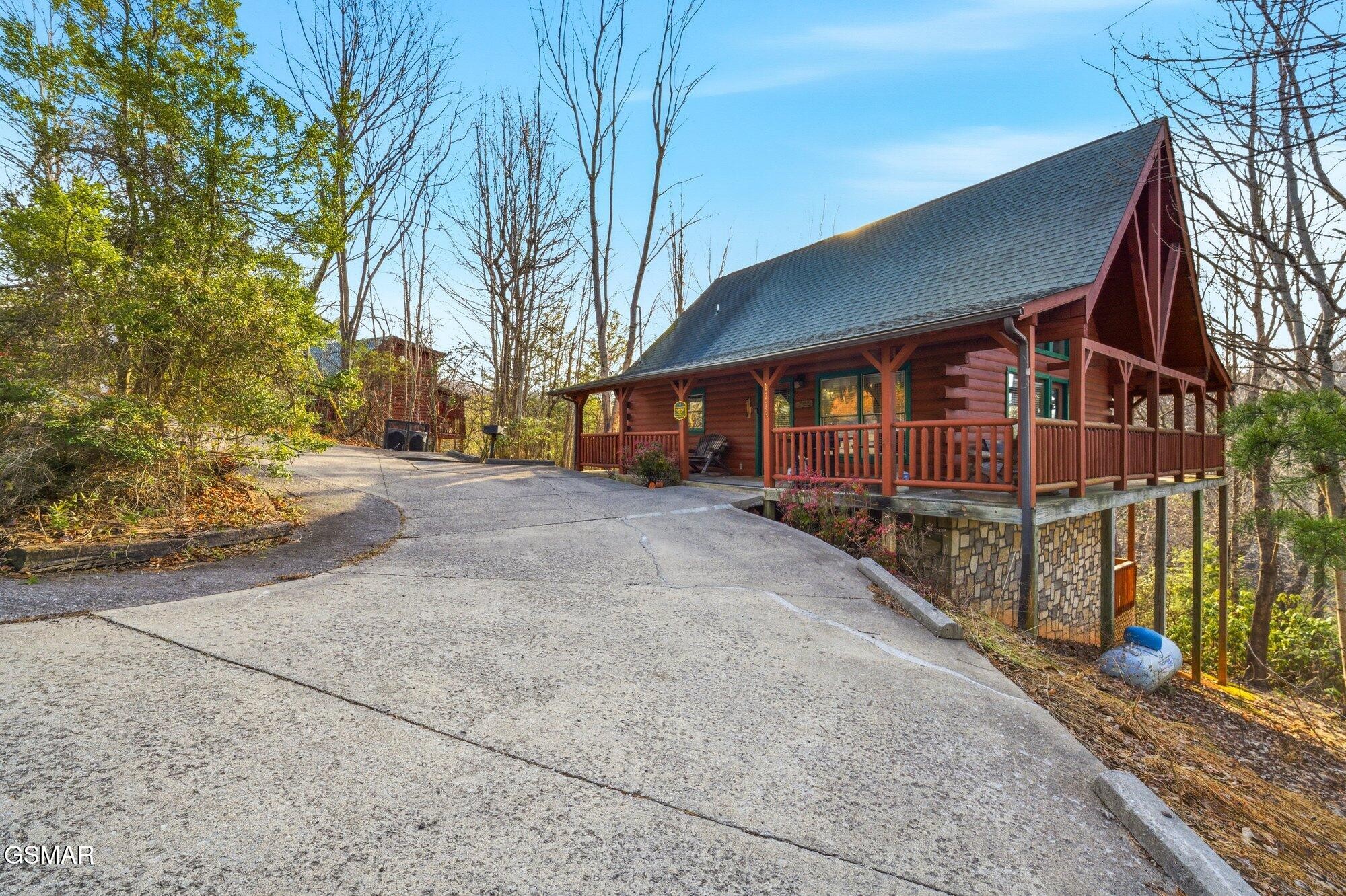 Log cabin with a covered wraparound porch and stone foundation beside a wooded driveway.