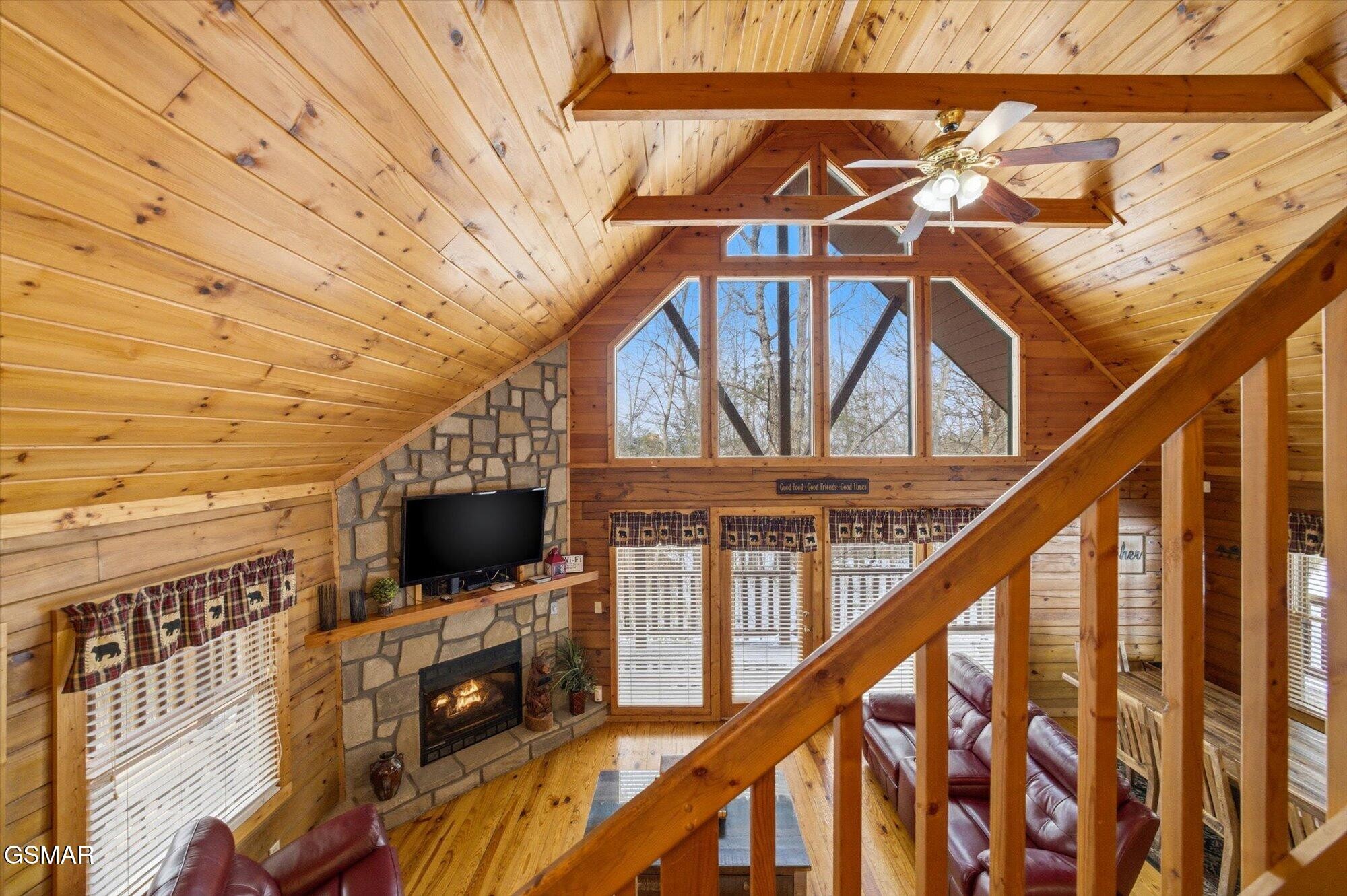 Cabin living room with vaulted pine ceiling, stone fireplace, tall windows, and wooden staircase railing.
