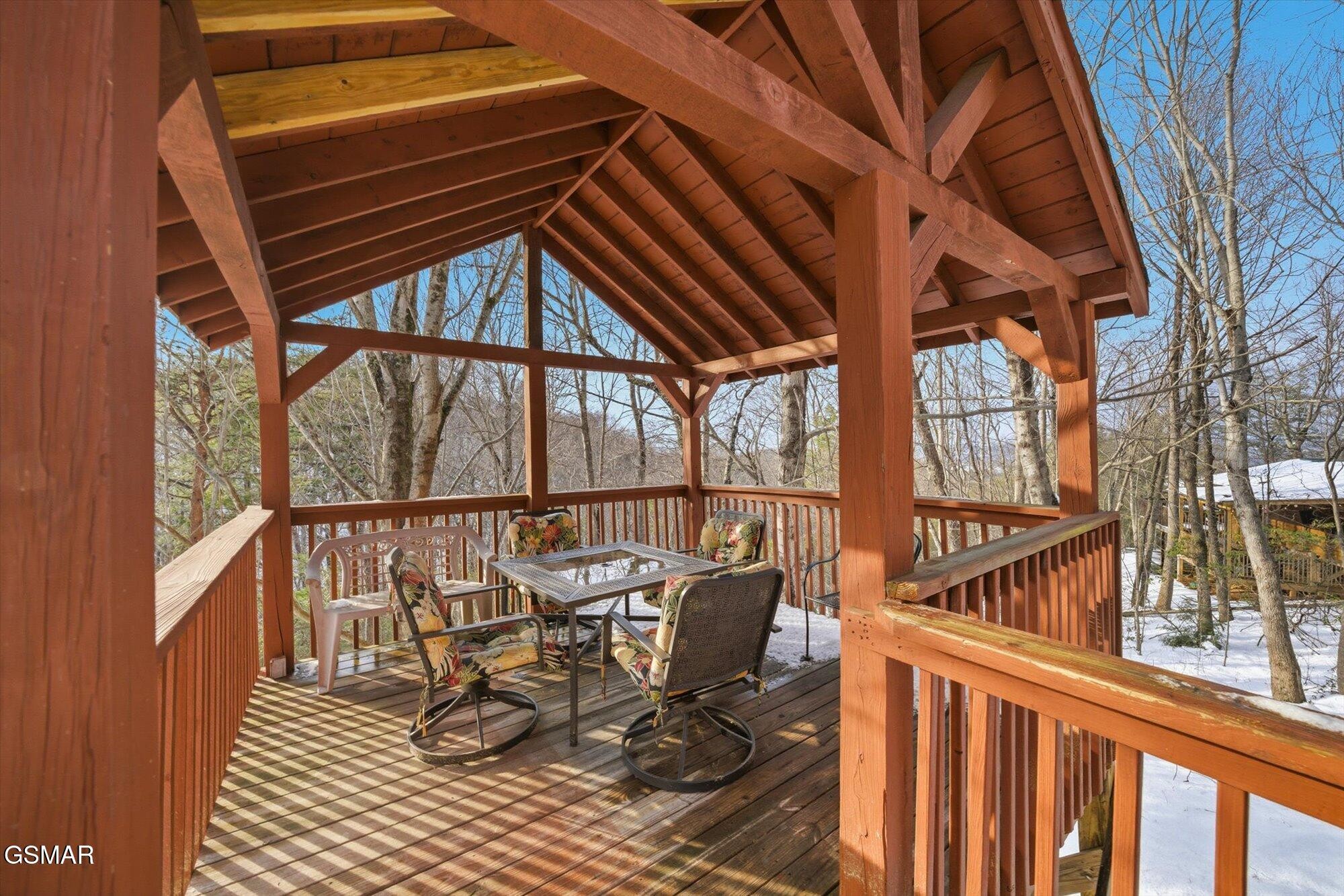 Covered wooden deck with timber beams, floral-cushioned chairs, and snowy woods beyond the railing.
