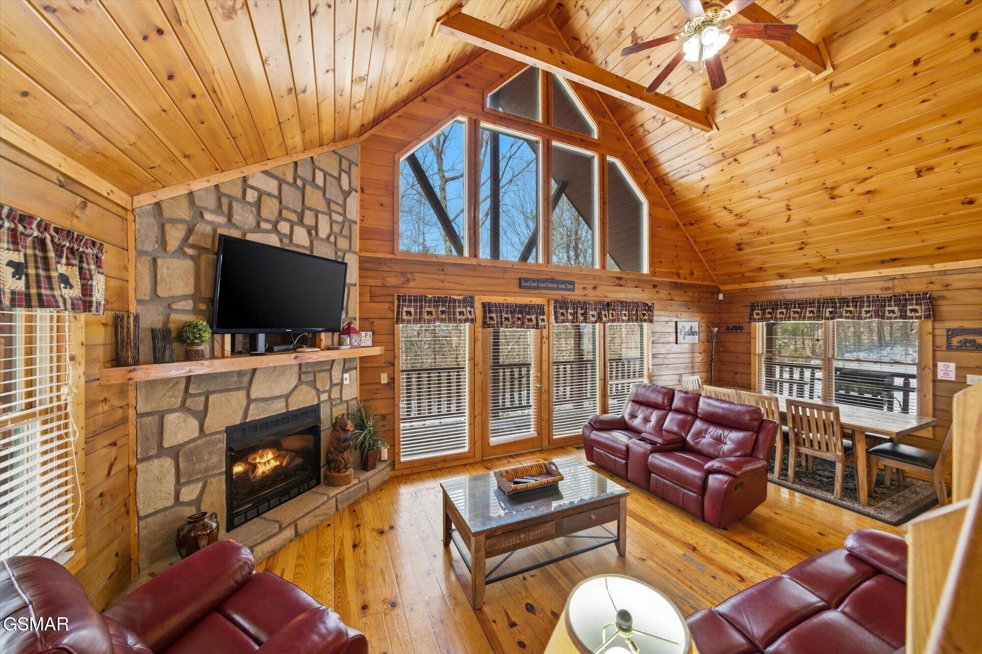 Cabin living room with vaulted pine ceiling, stone fireplace, tall windows, and burgundy leather sofas.