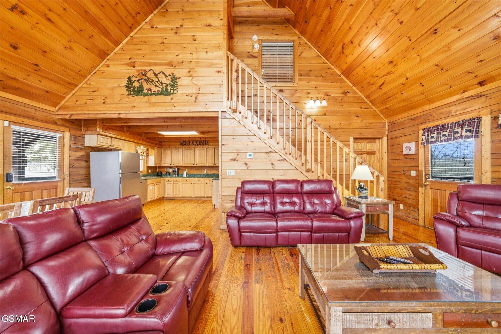 Cabin living room with vaulted pine ceiling, burgundy leather seating, staircase, and open kitchen.