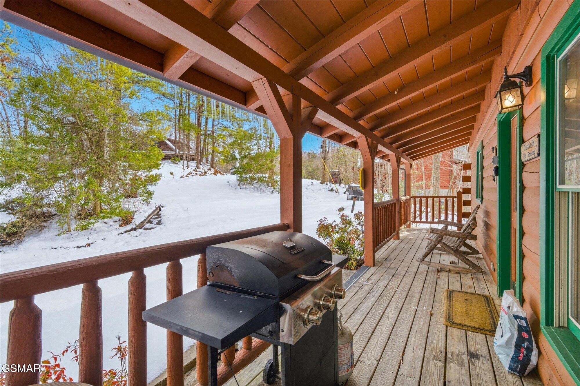 Covered cabin porch with a barbecue grill and rocking chairs overlooking a snowy wooded yard.