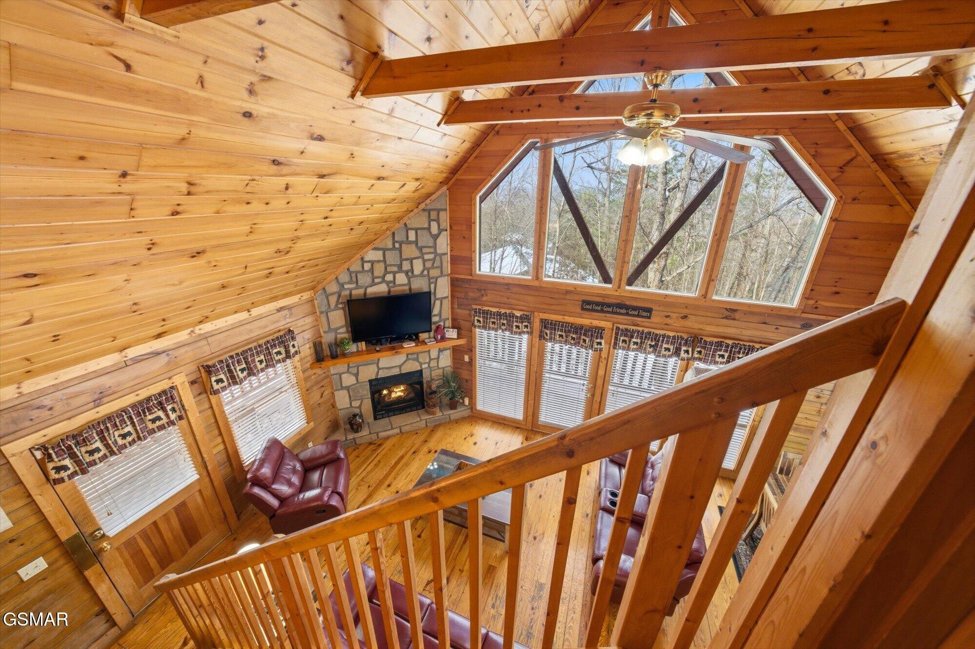Cabin living room with vaulted wood ceiling, large windows, and a stone fireplace, viewed from the loft.