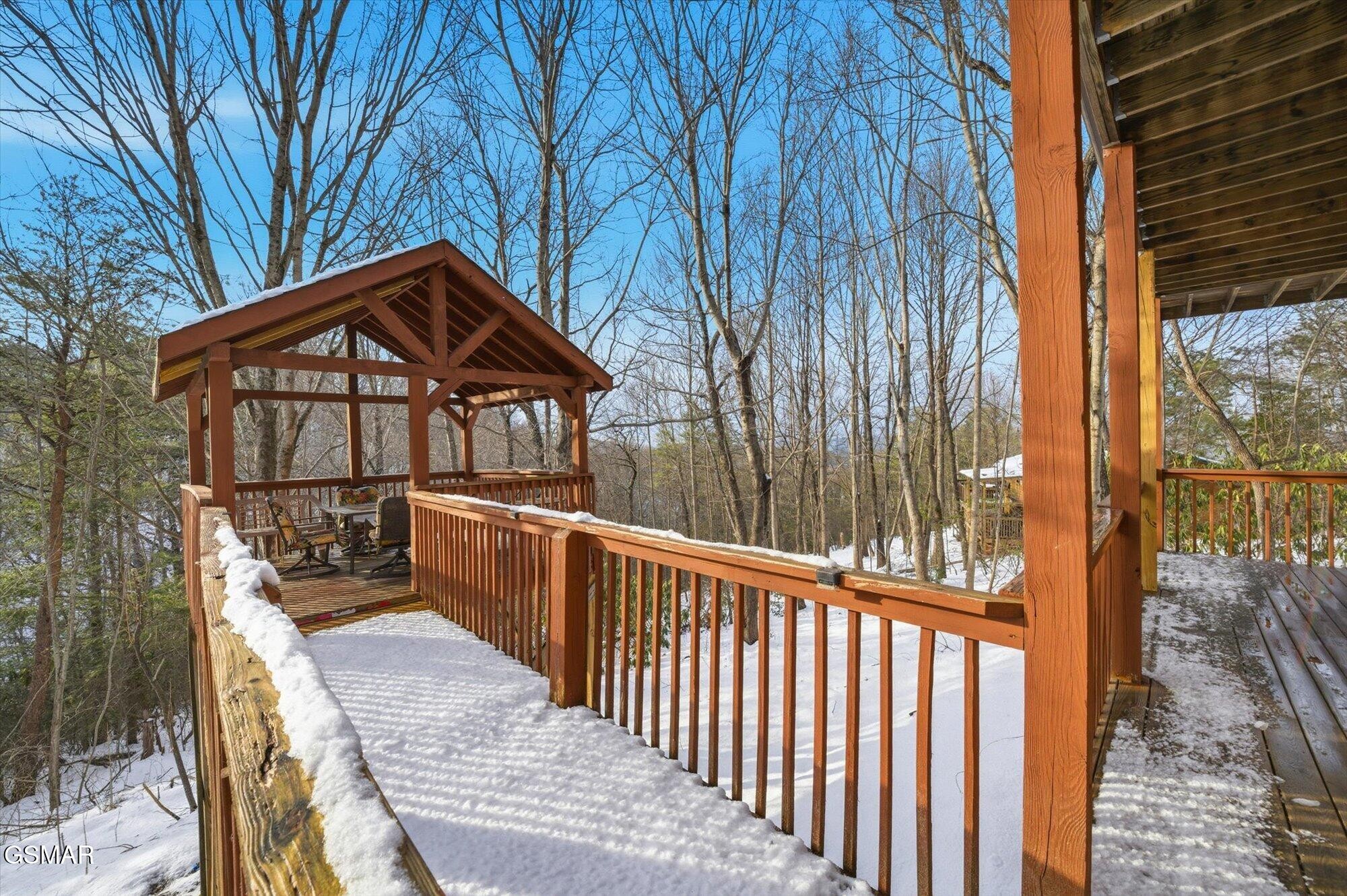 Snow-covered wooden deck leads to a covered gazebo with seating among leafless trees.