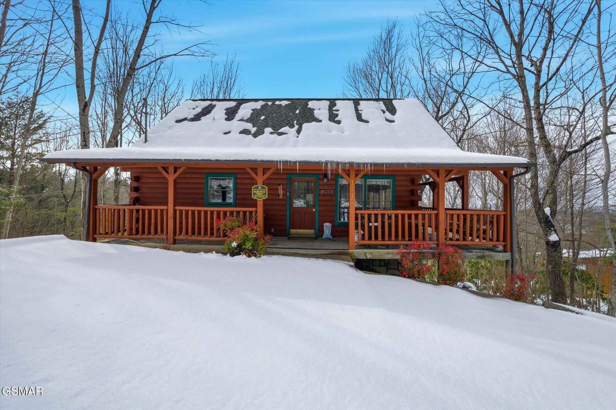 Log cabin with a wide covered porch, snow-covered roof and yard, and icicles along the eaves.