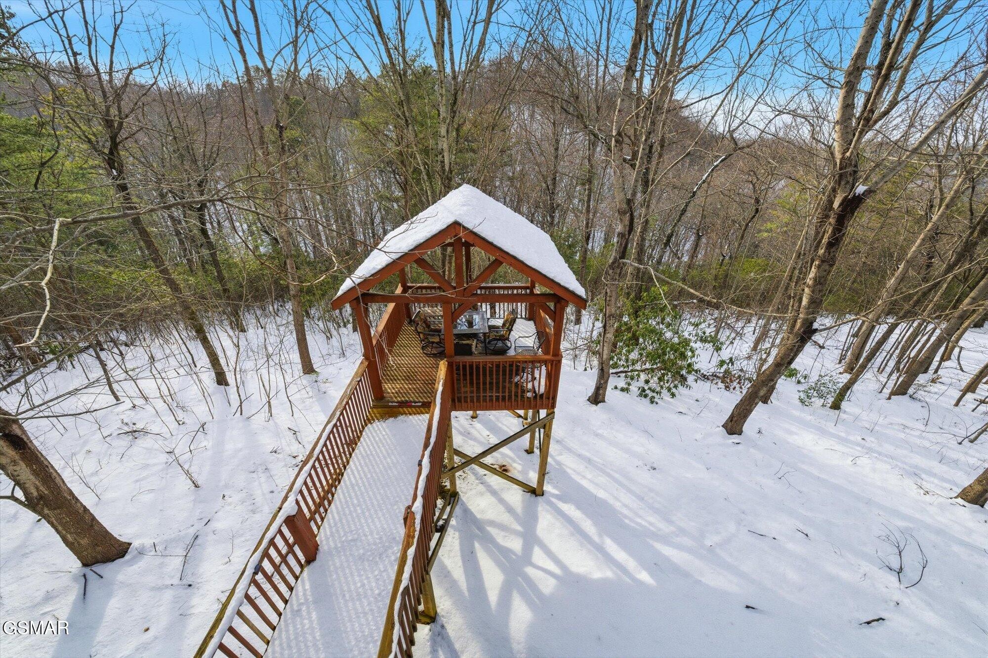 Raised wooden gazebo with a snow-covered roof and railed walkway in a snowy woodland.