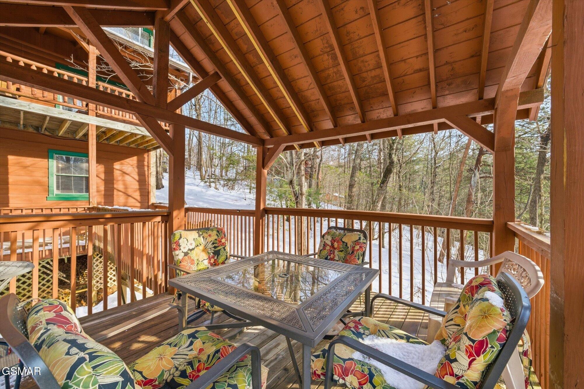 Covered wooden deck with floral patio chairs and a glass-top table, overlooking a snowy wooded hillside.