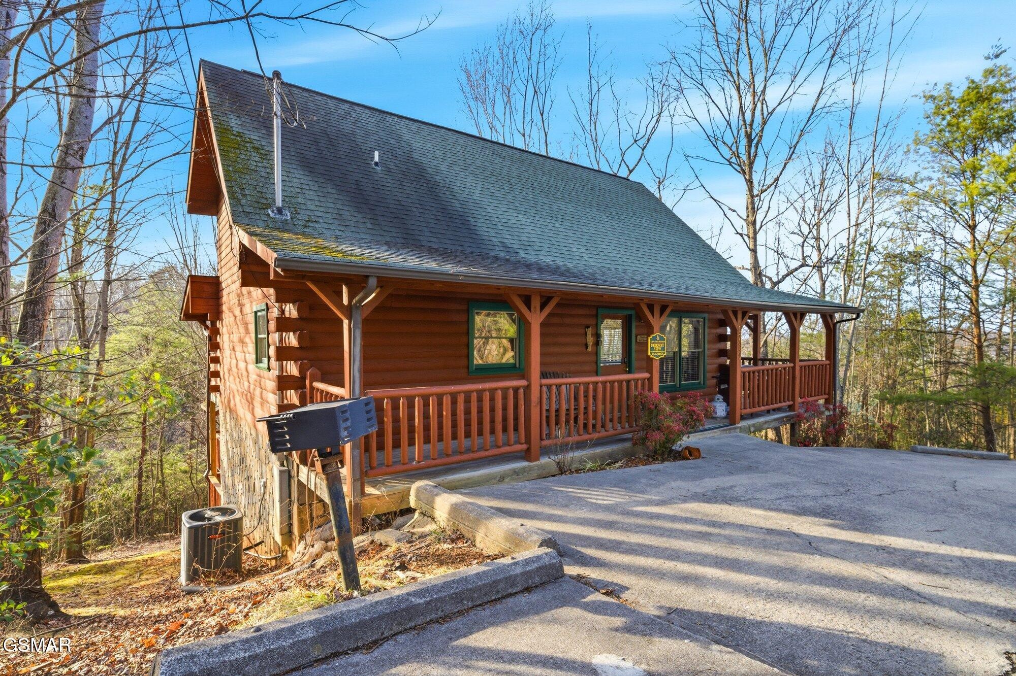 Log cabin with a covered porch beside a paved driveway, surrounded by trees.