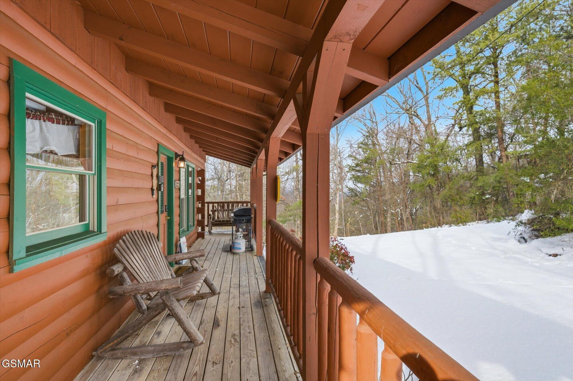 Covered log cabin porch with rustic chairs and a grill beside a snowy wooded yard.