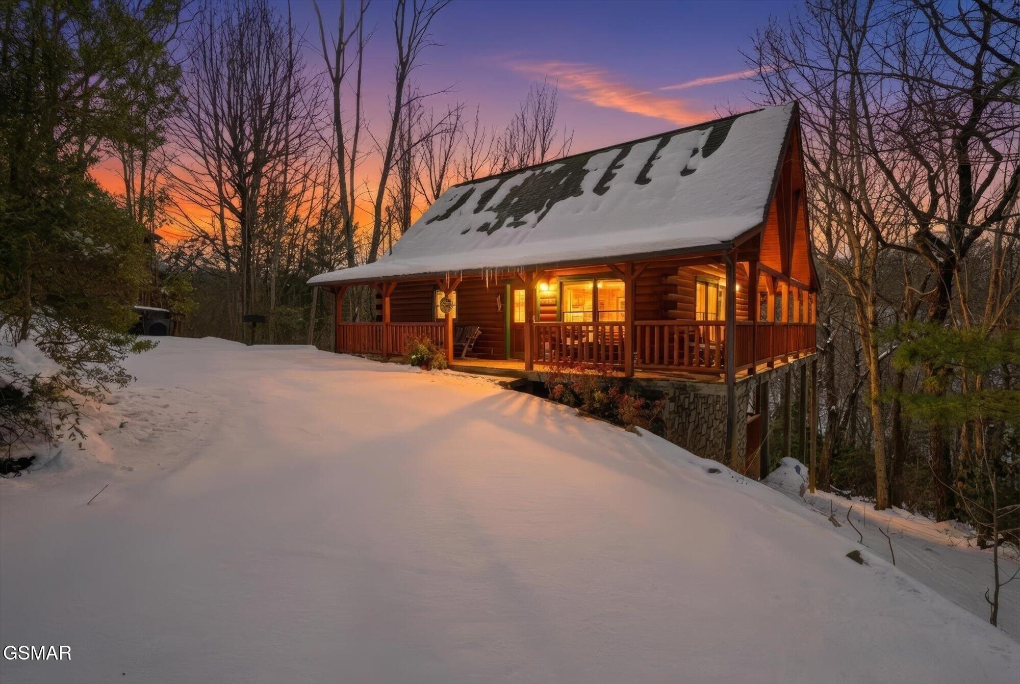 Snow-covered log cabin glowing warmly at dusk, with a wraparound porch and wooded surroundings.