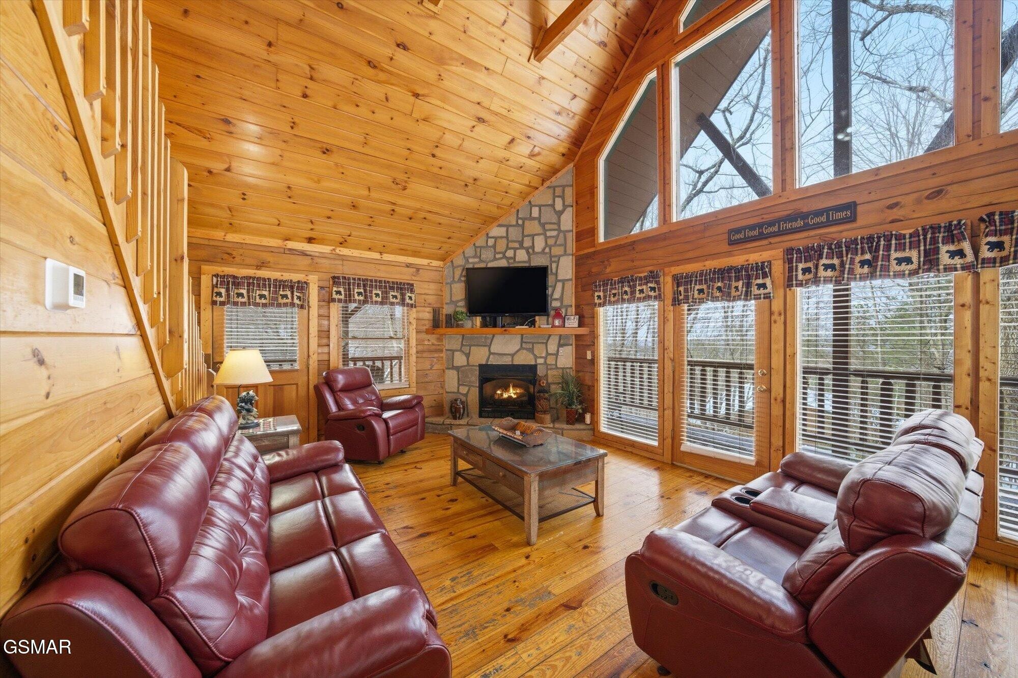 Cabin living room with vaulted wood ceilings, a stone fireplace, burgundy leather seating, and tall windows.