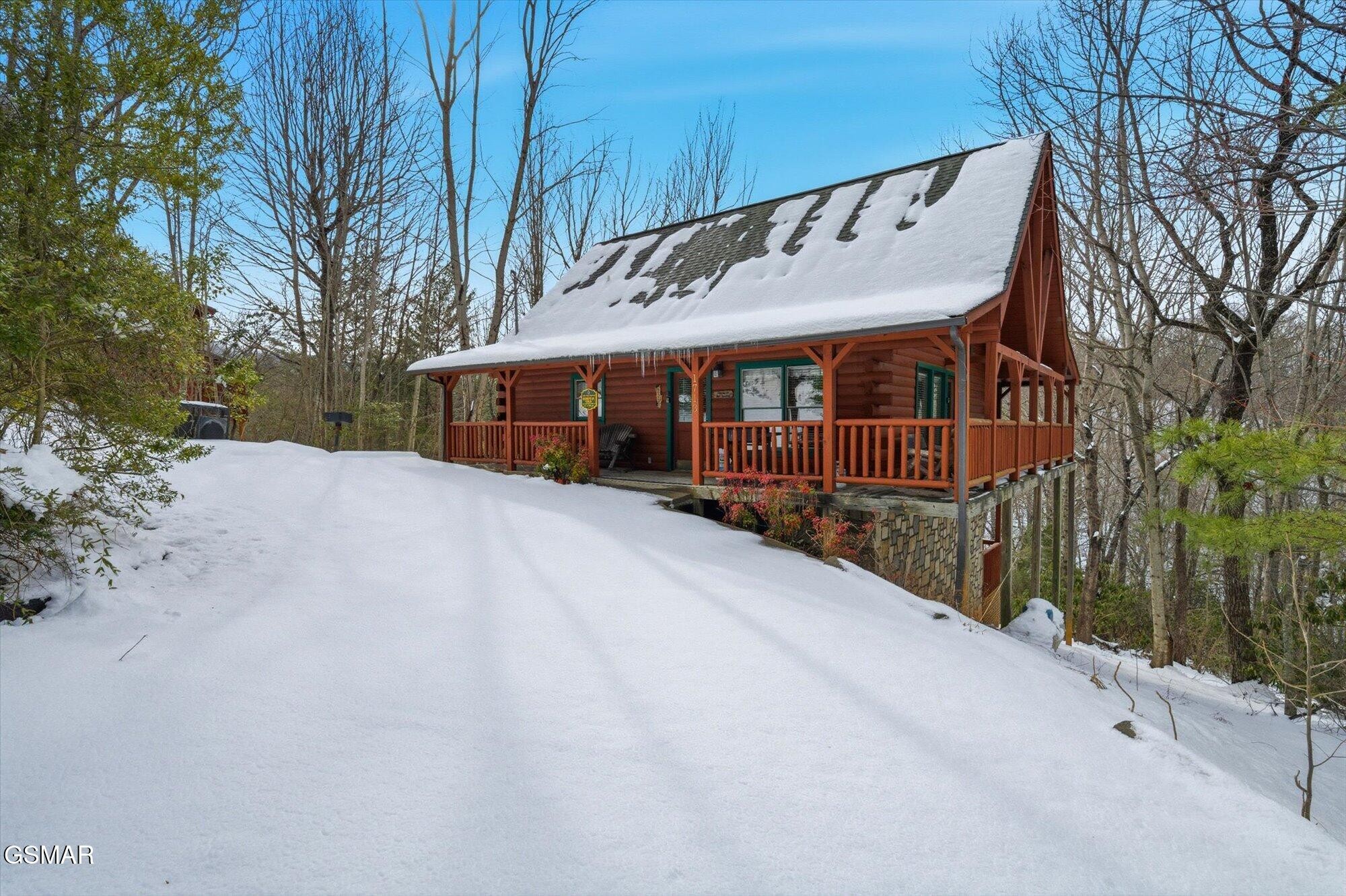Log cabin with a wraparound porch on a snowy wooded slope.