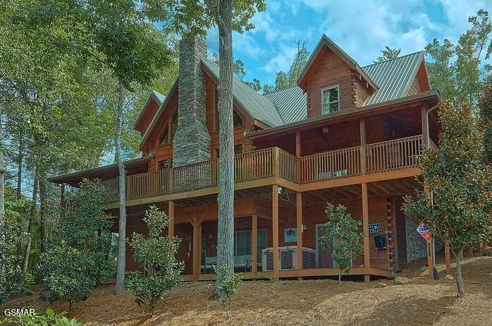 Two-story wood cabin with a wraparound porch and stone chimney, surrounded by trees.