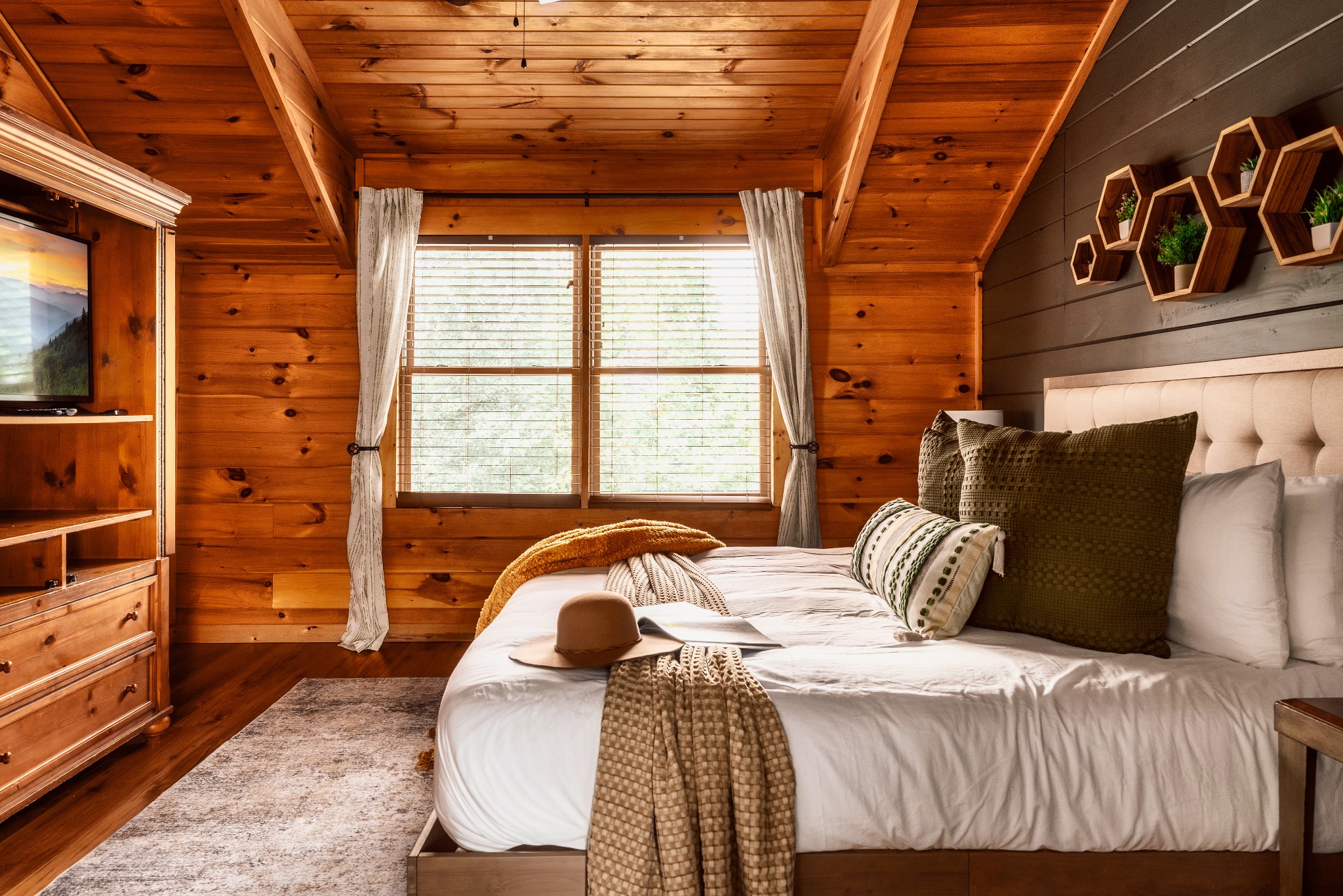 Cabin bedroom with a white bed, vaulted wood ceiling, and large curtained window.