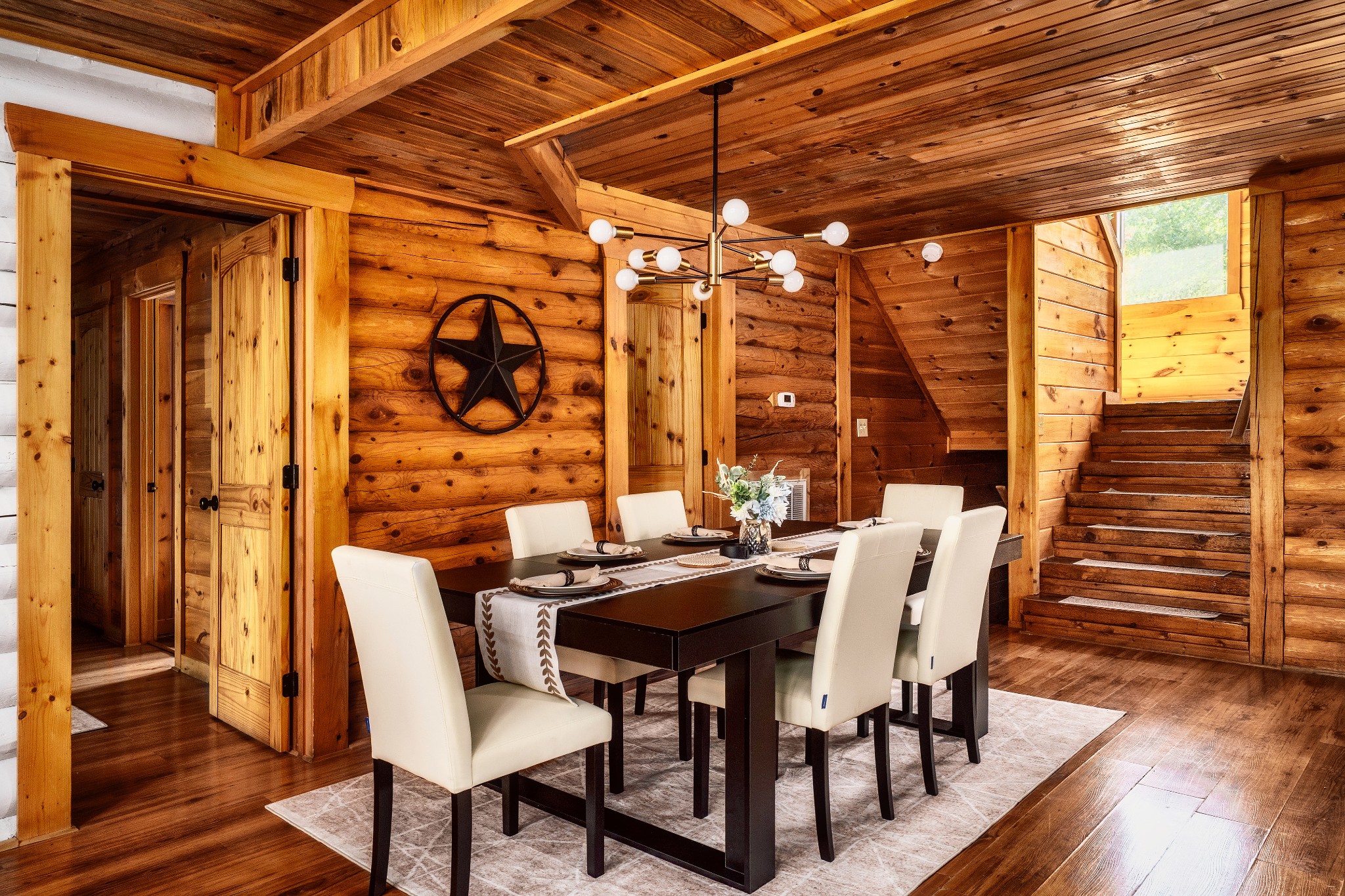 Cabin dining room with a set table, white chairs, log walls, and a staircase.