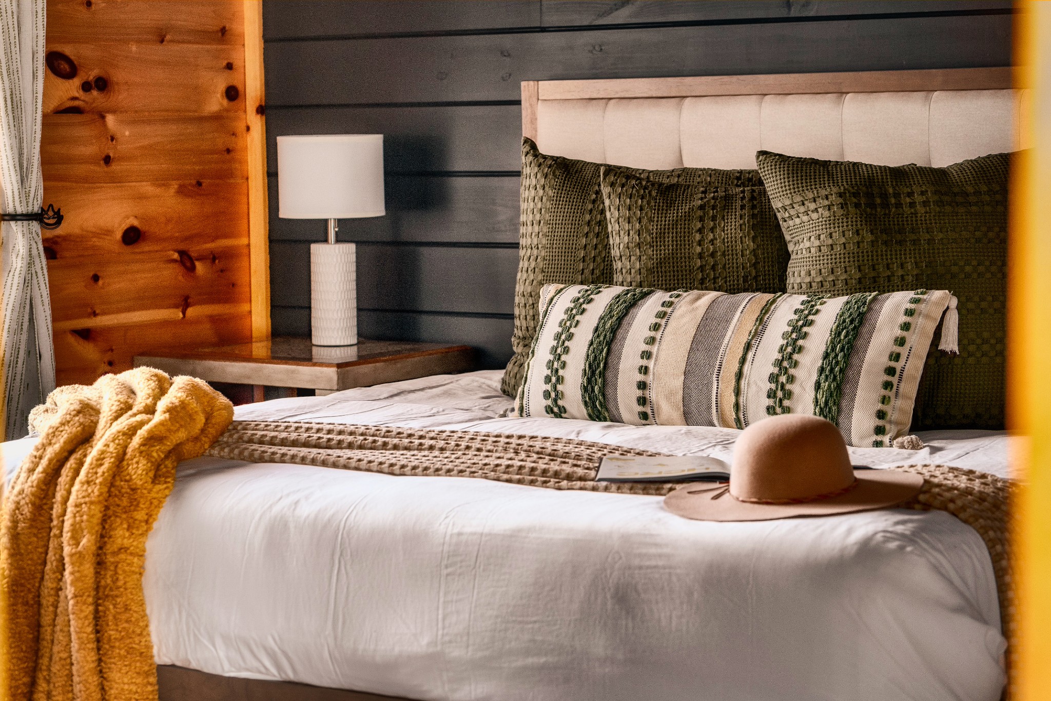 Bedroom with green and cream pillows, a mustard knit throw, and a tan hat on the bed.