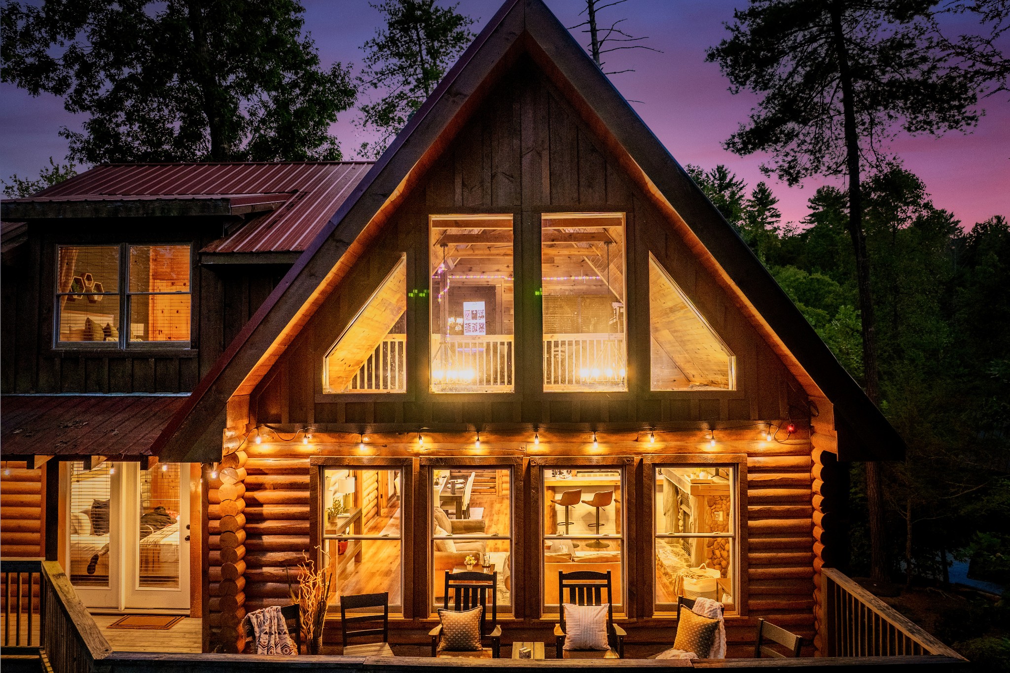 Log cabin exterior at dusk, warmly lit windows beneath a steep A-frame roof and deck seating.