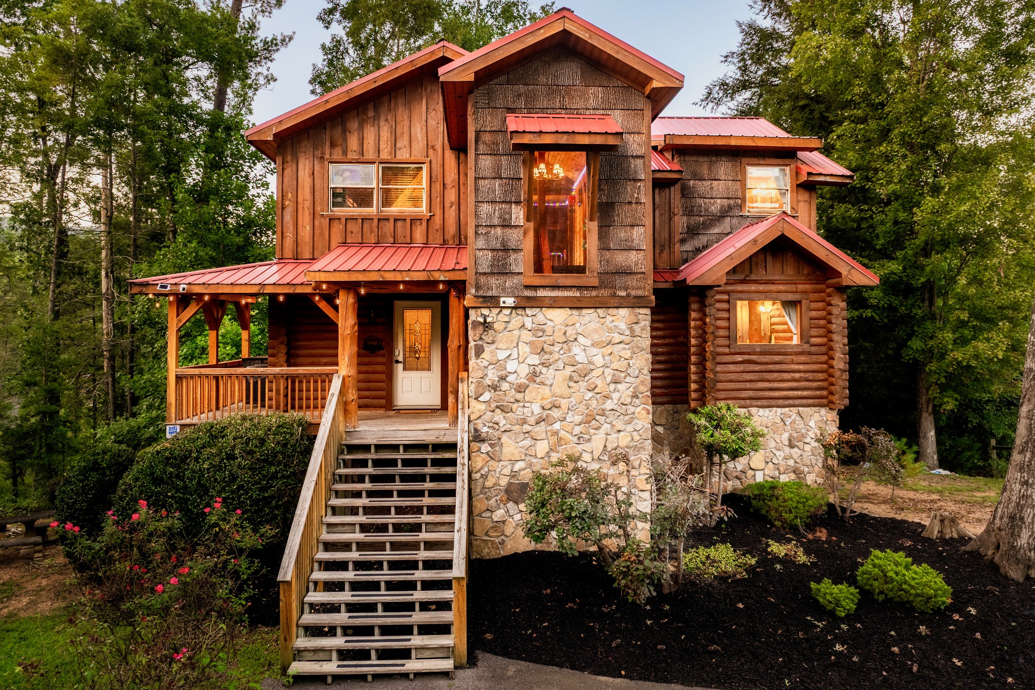 Rustic cabin with stone and log siding, red metal roofs, and a covered porch among trees.