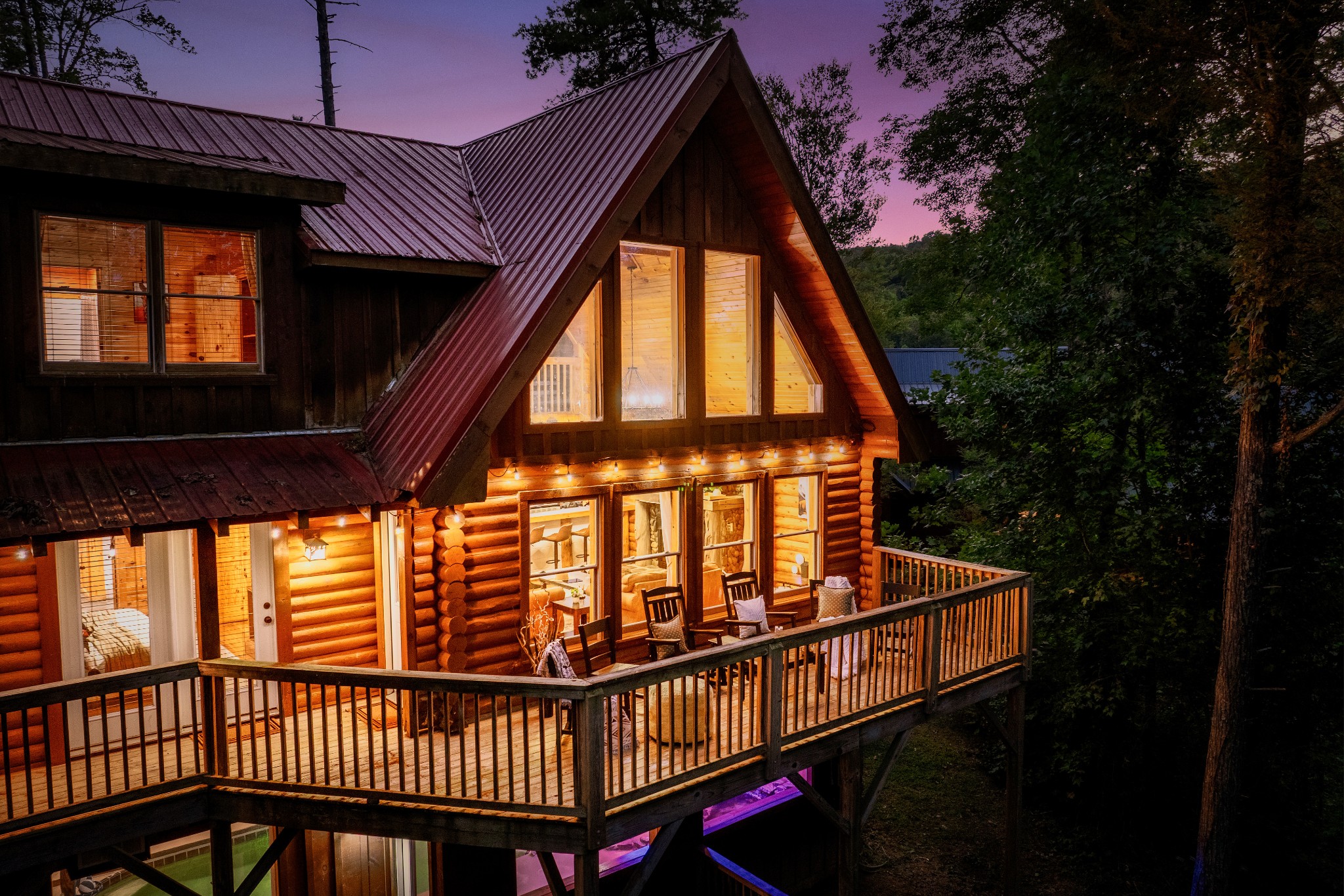 Log cabin at dusk with a steep metal roof and a raised deck among trees.