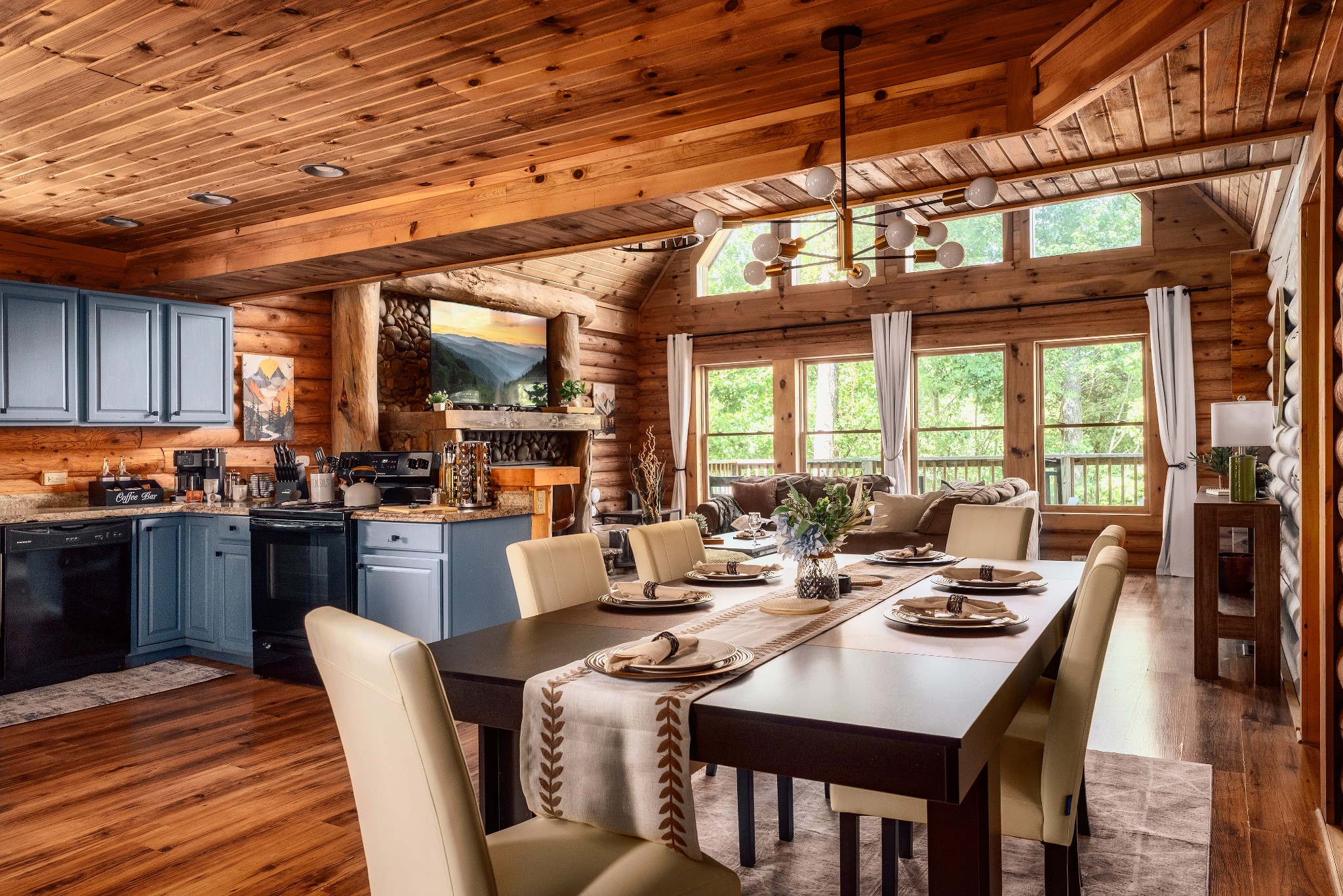 Open-plan cabin dining area and kitchen with a set table, blue cabinets, stone fireplace, and vaulted timber ceiling.