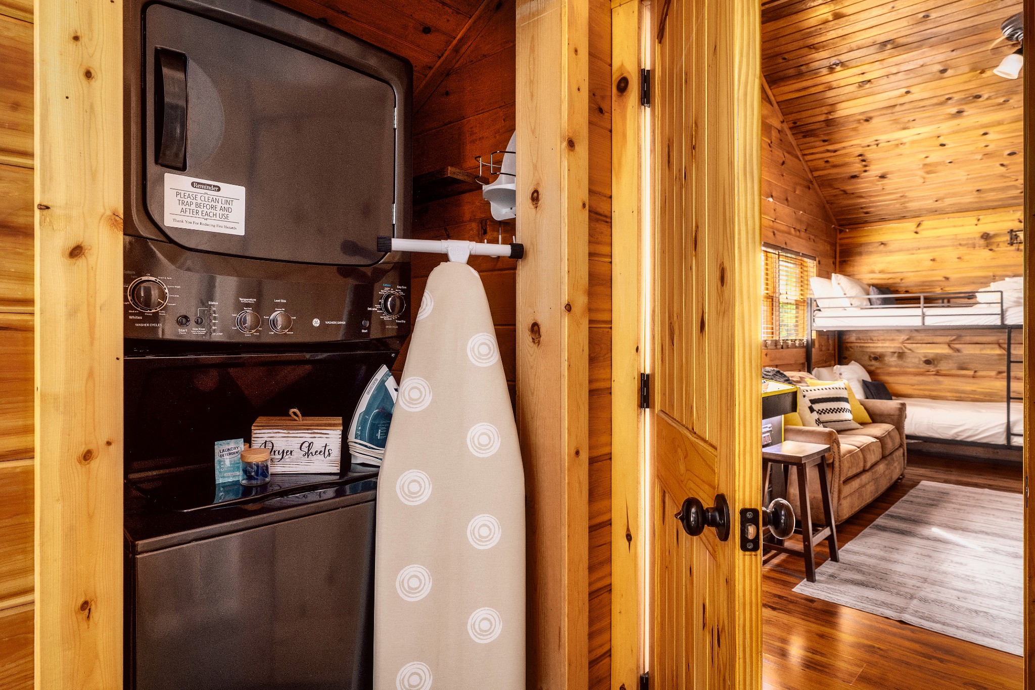 Cabin laundry area with stacked washer and dryer, ironing board, and a bunk bedroom visible through the doorway.