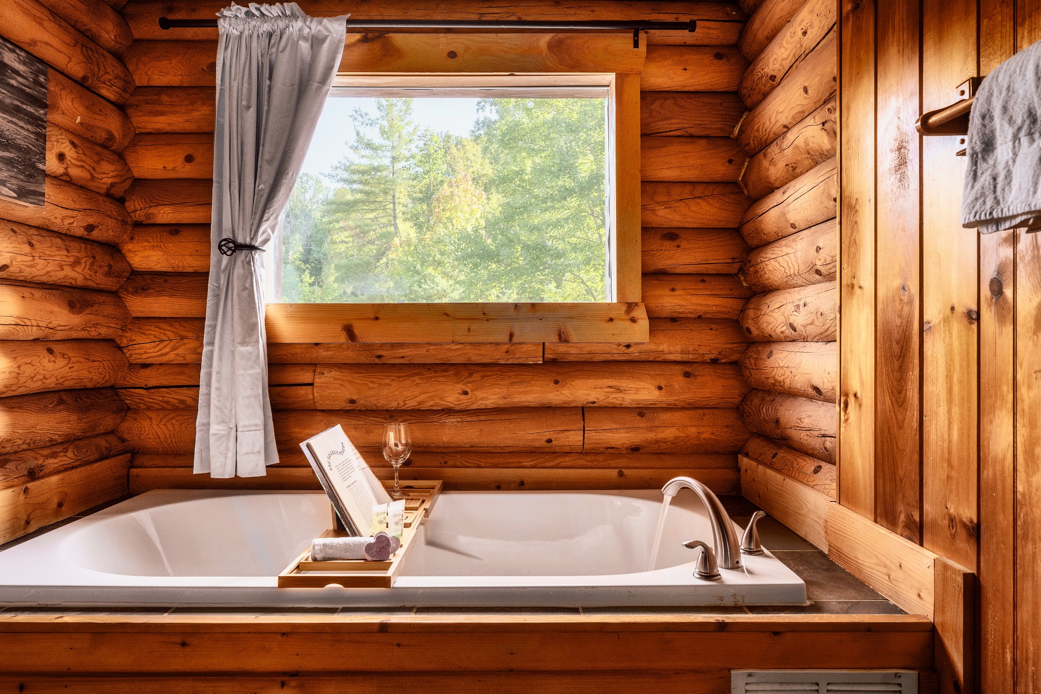 Soaking tub in a log cabin bathroom, with a window overlooking leafy trees.