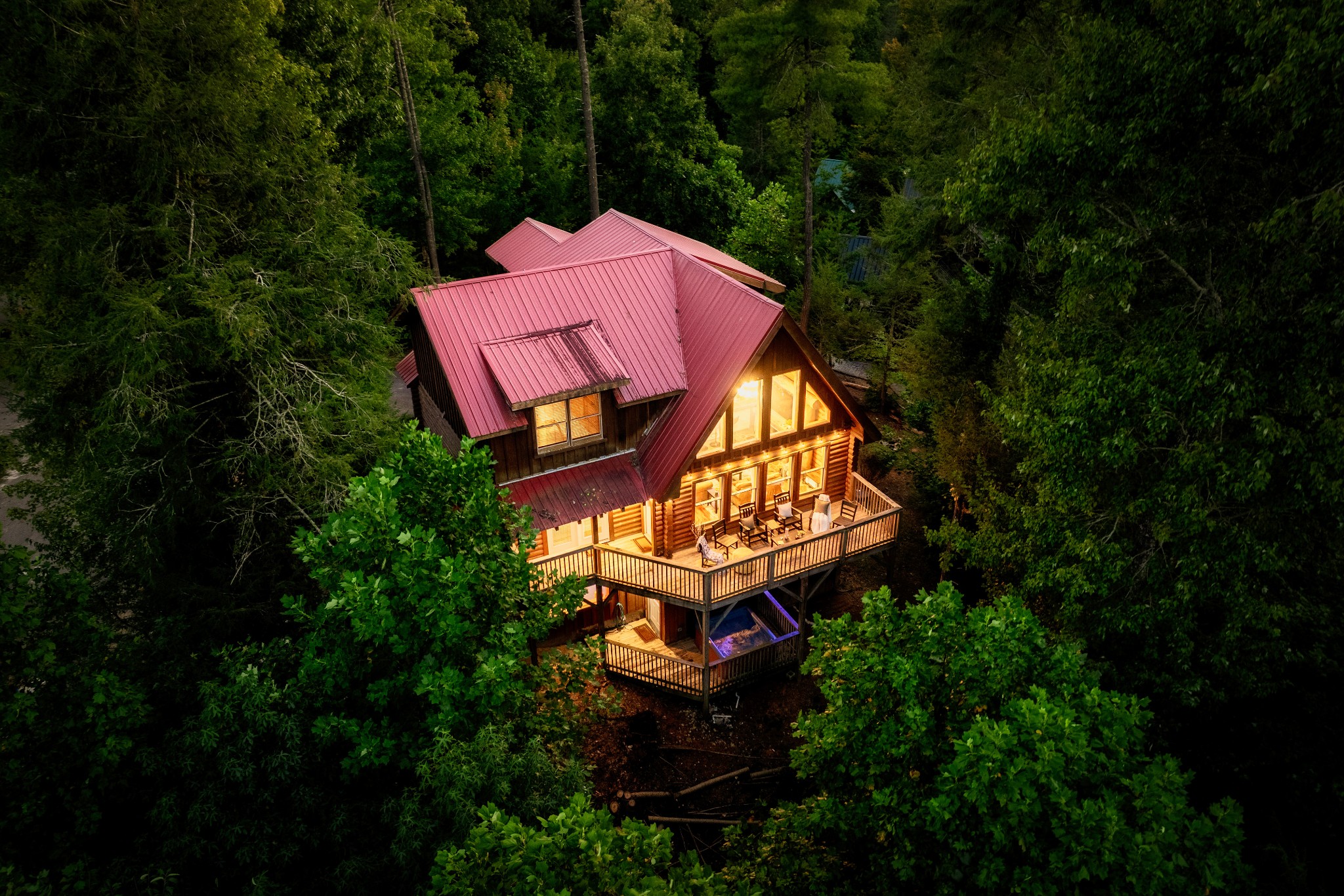 Aerial view of a log cabin with a red metal roof and warmly lit decks among dense trees at night.