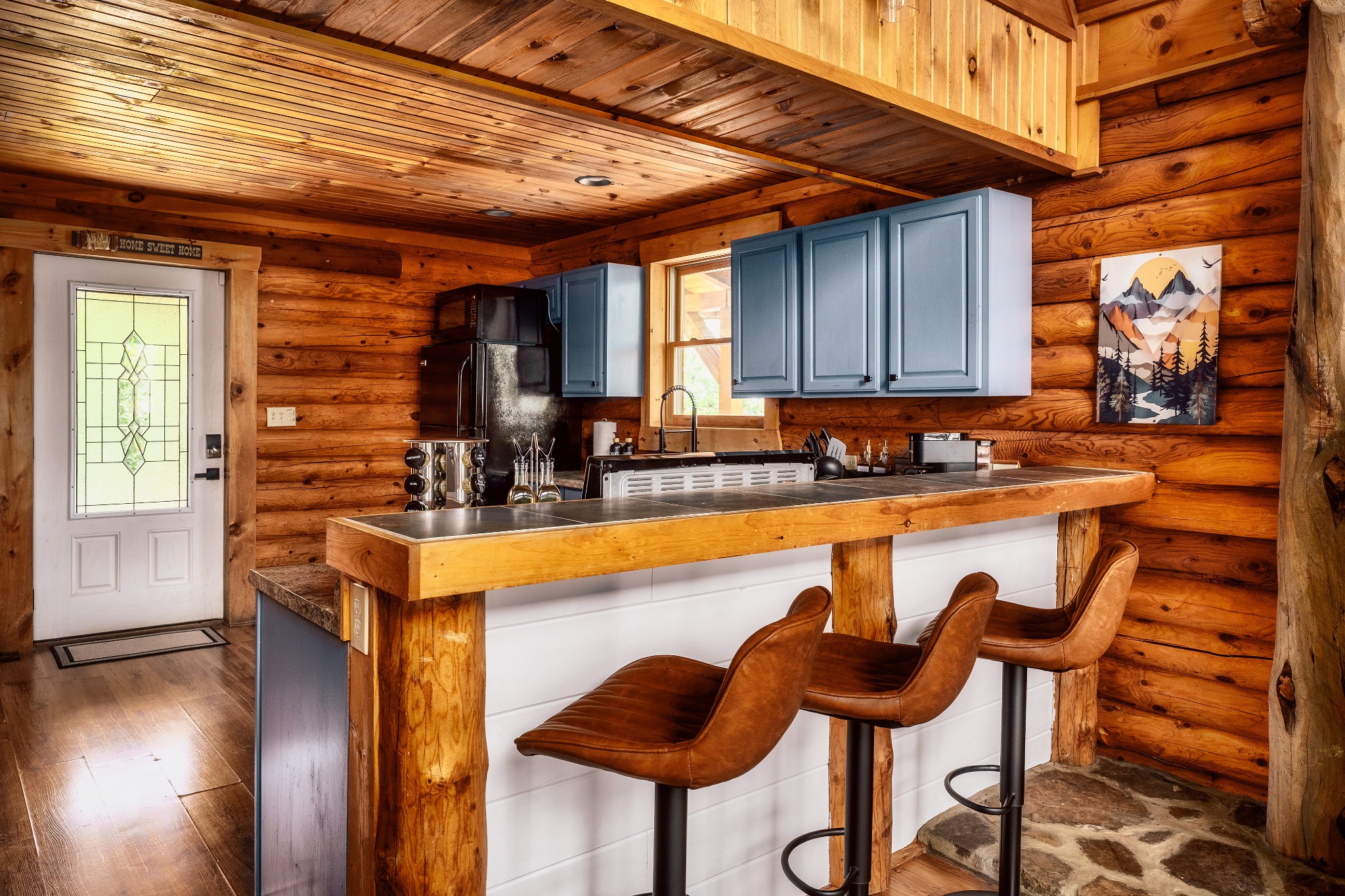 Rustic log cabin kitchen with blue-gray cabinets and a wood-trimmed island lined with leather bar stools.