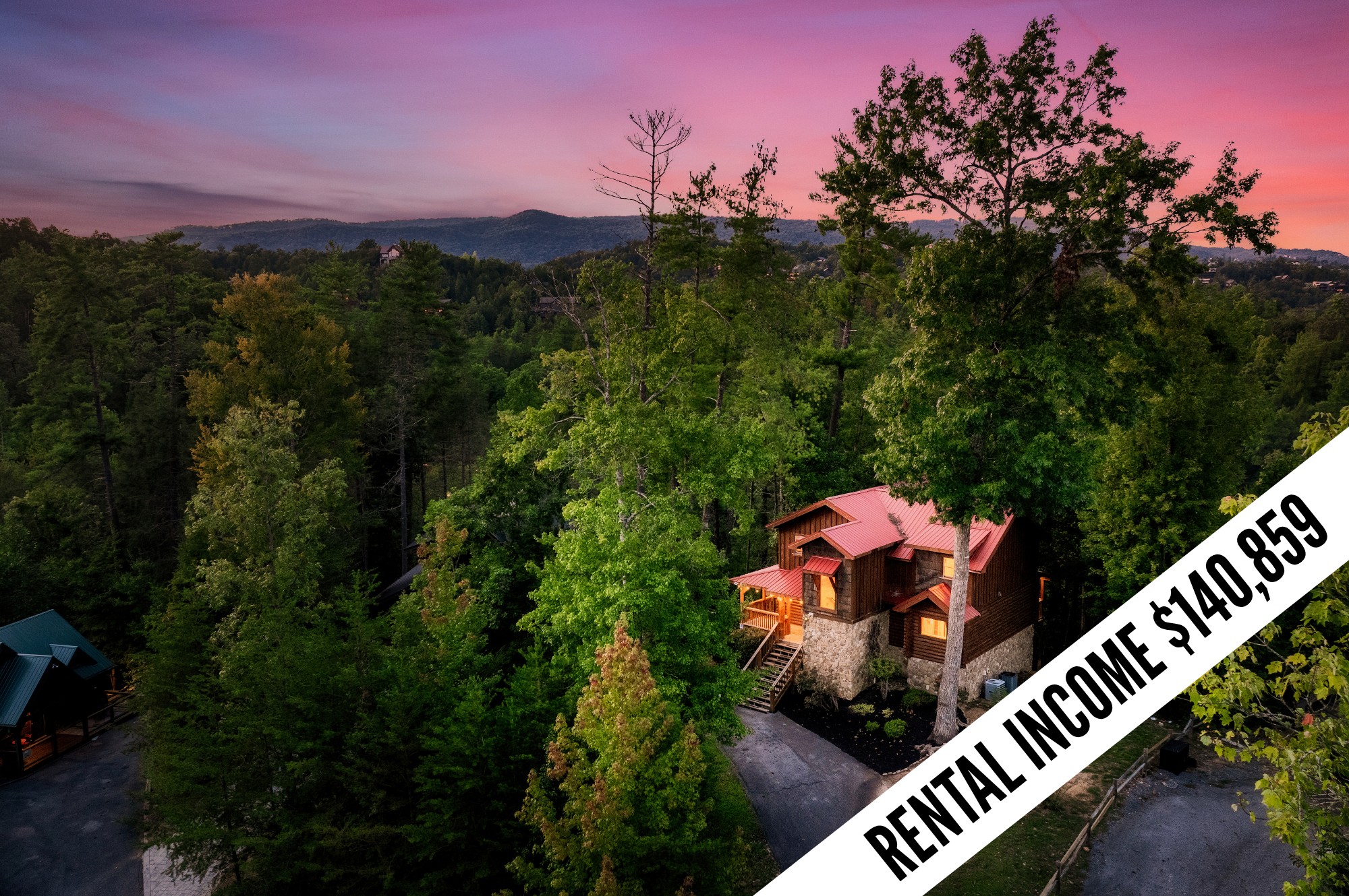 Aerial view of a stone-and-wood cabin among dense trees, with a pink dusk sky and distant mountain ridges.