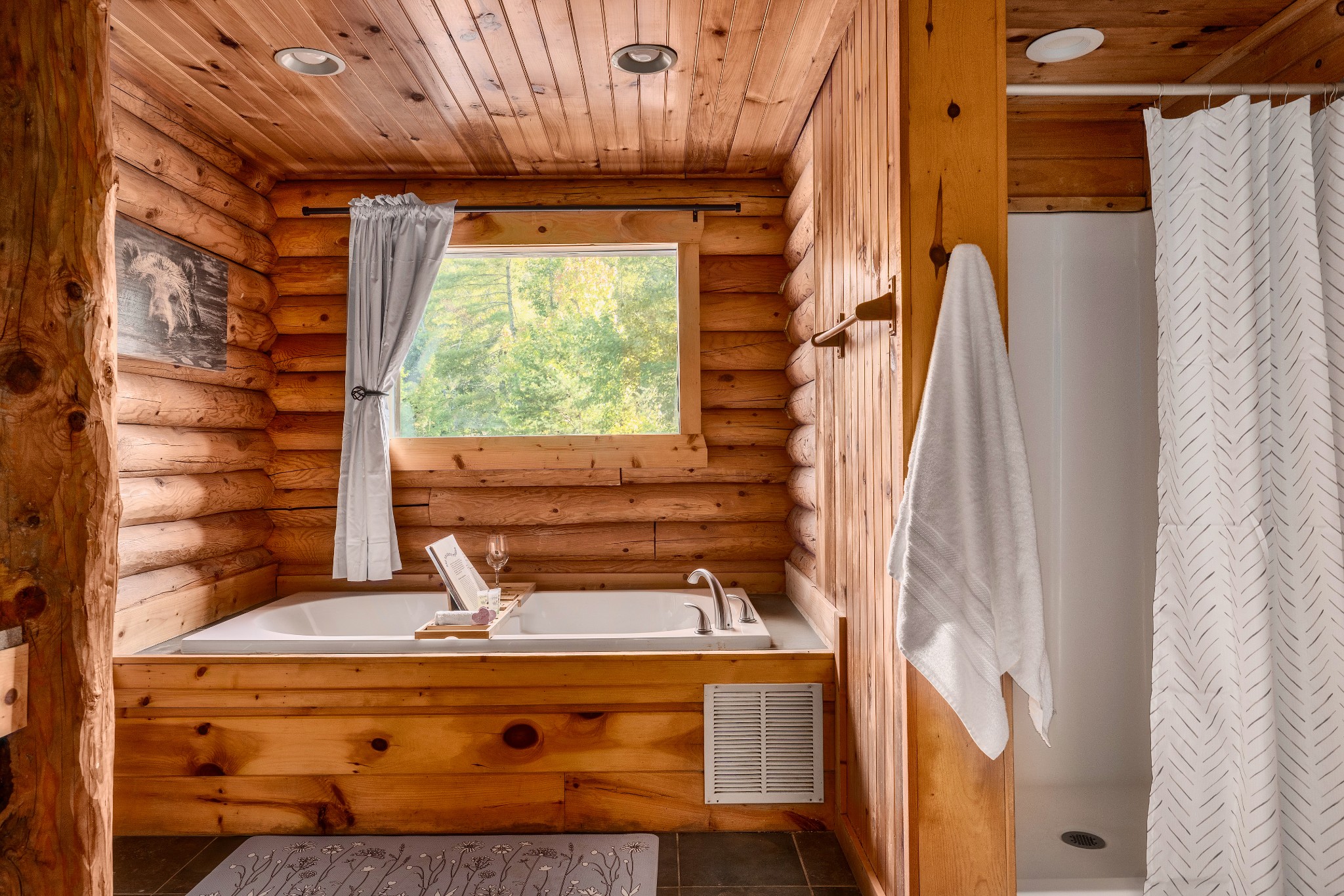 Log-cabin bathroom with a soaking tub beneath a leafy window beside a curtained shower.