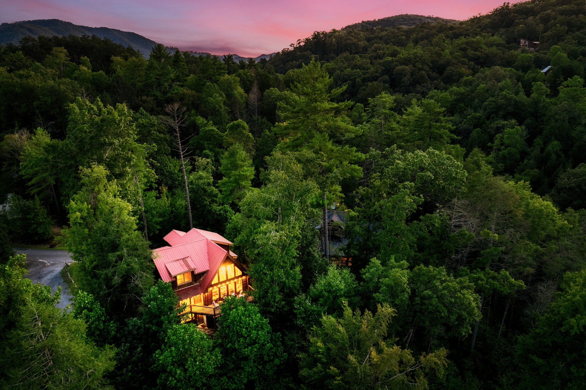 Aerial view of a red-roofed log cabin glowing among dense forest and mountain ridges at dusk.