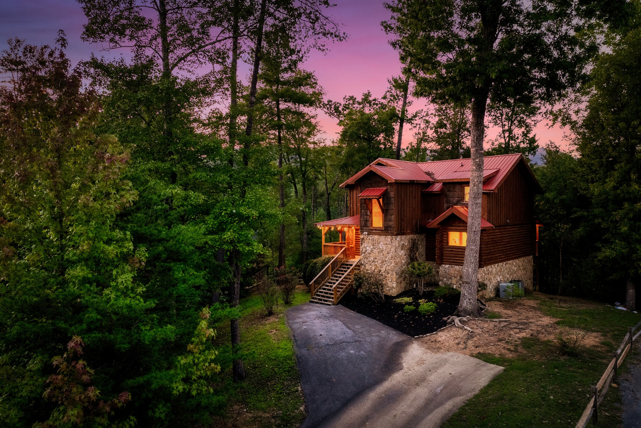 Rustic stone-and-log cabin with a red roof and glowing porch, surrounded by trees at dusk.