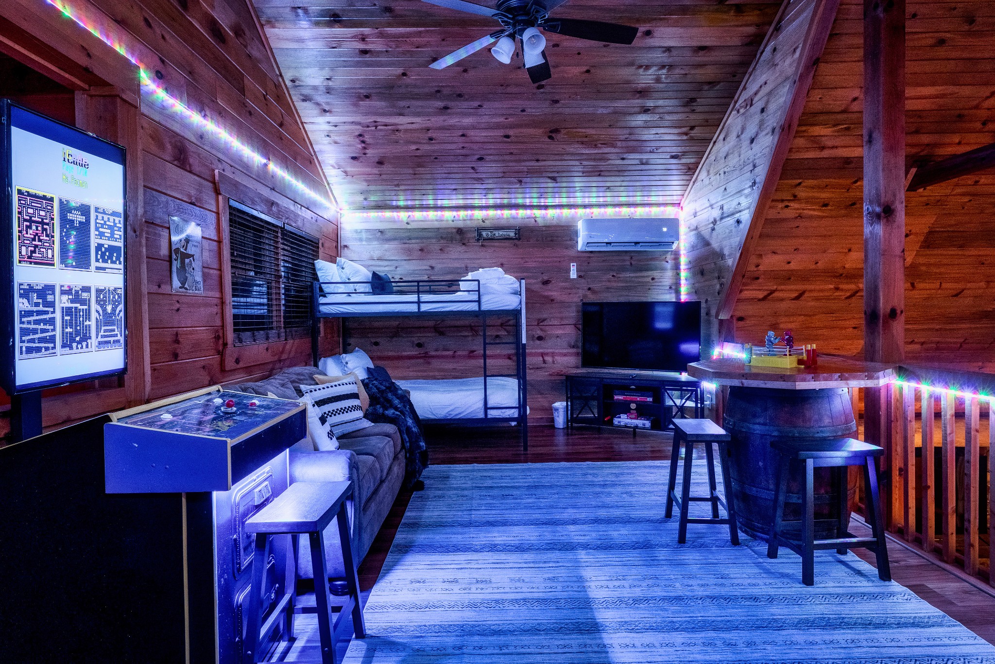 Wood-paneled cabin loft game room with a bunk bed, arcade game, and colorful string lights.