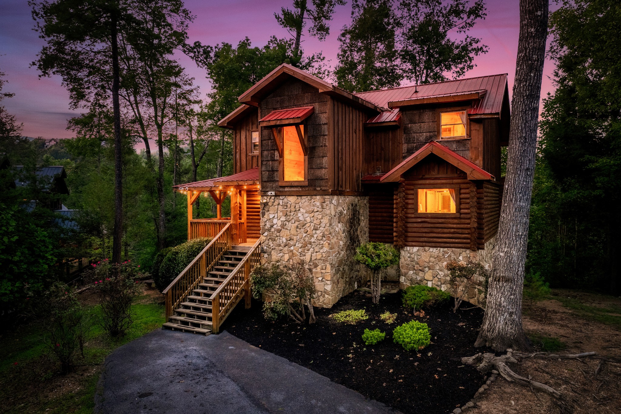 Rustic cabin with a stone foundation and glowing windows, viewed from the driveway at dusk.