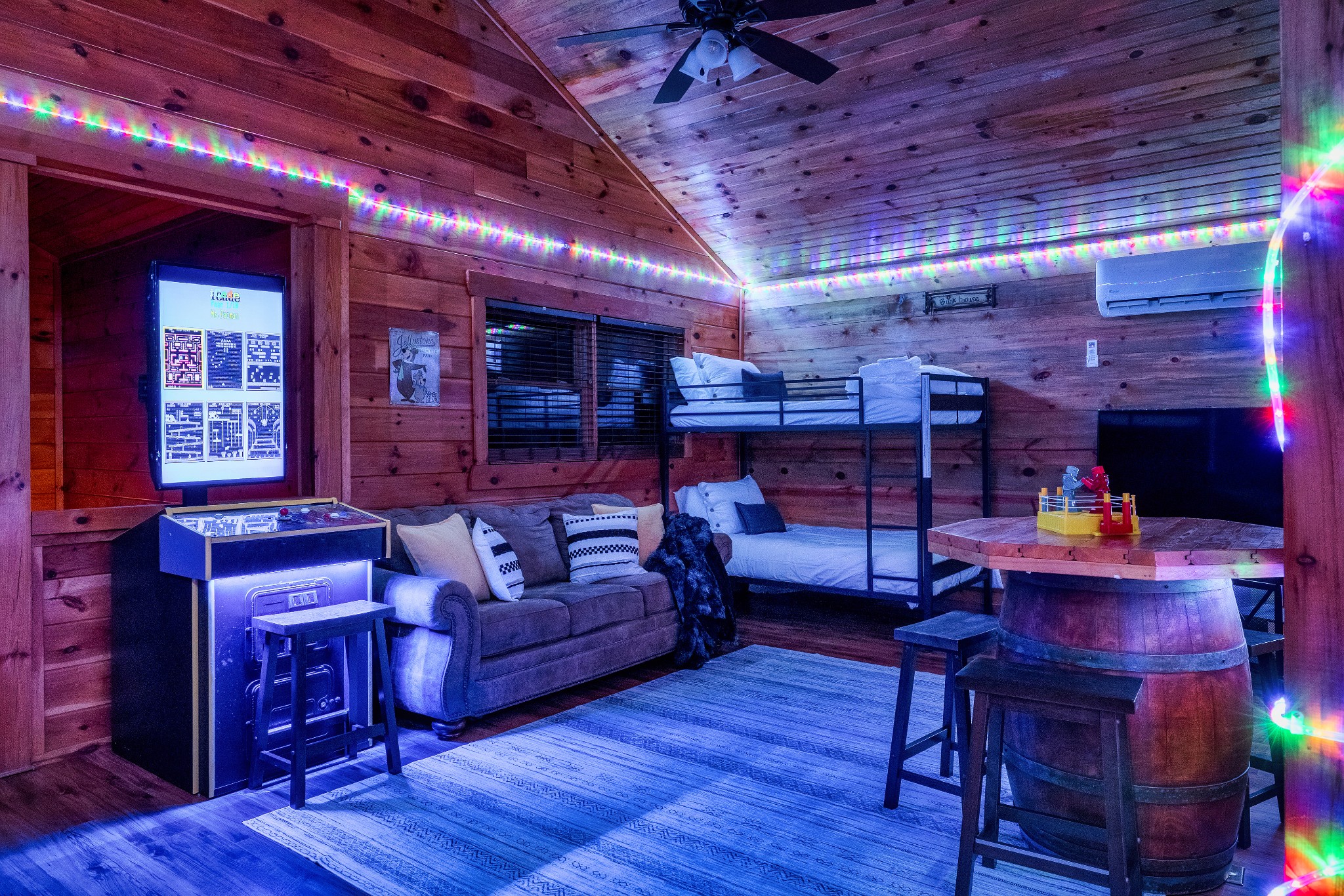 Wood-paneled cabin game room with bunk beds, arcade game, sofa, and colorful lights.