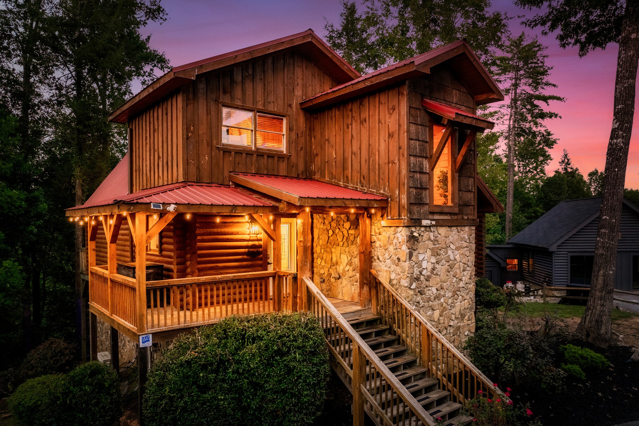 Rustic wood-and-stone cabin glowing at dusk, with a covered porch and long wooden staircase.