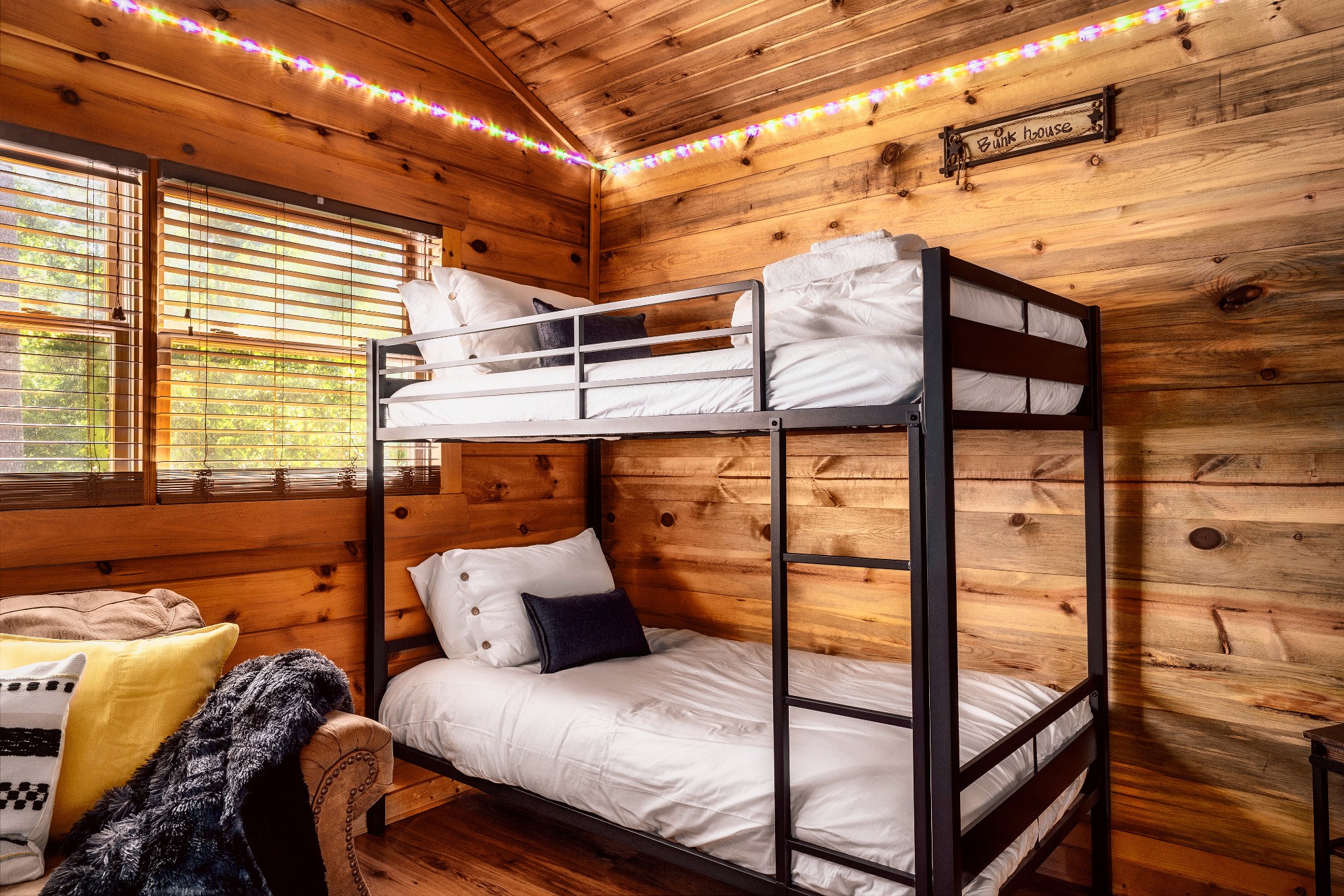 Wood-paneled bunk bedroom with a black metal bunk bed, colorful ceiling lights, and a “Bunk house” sign.