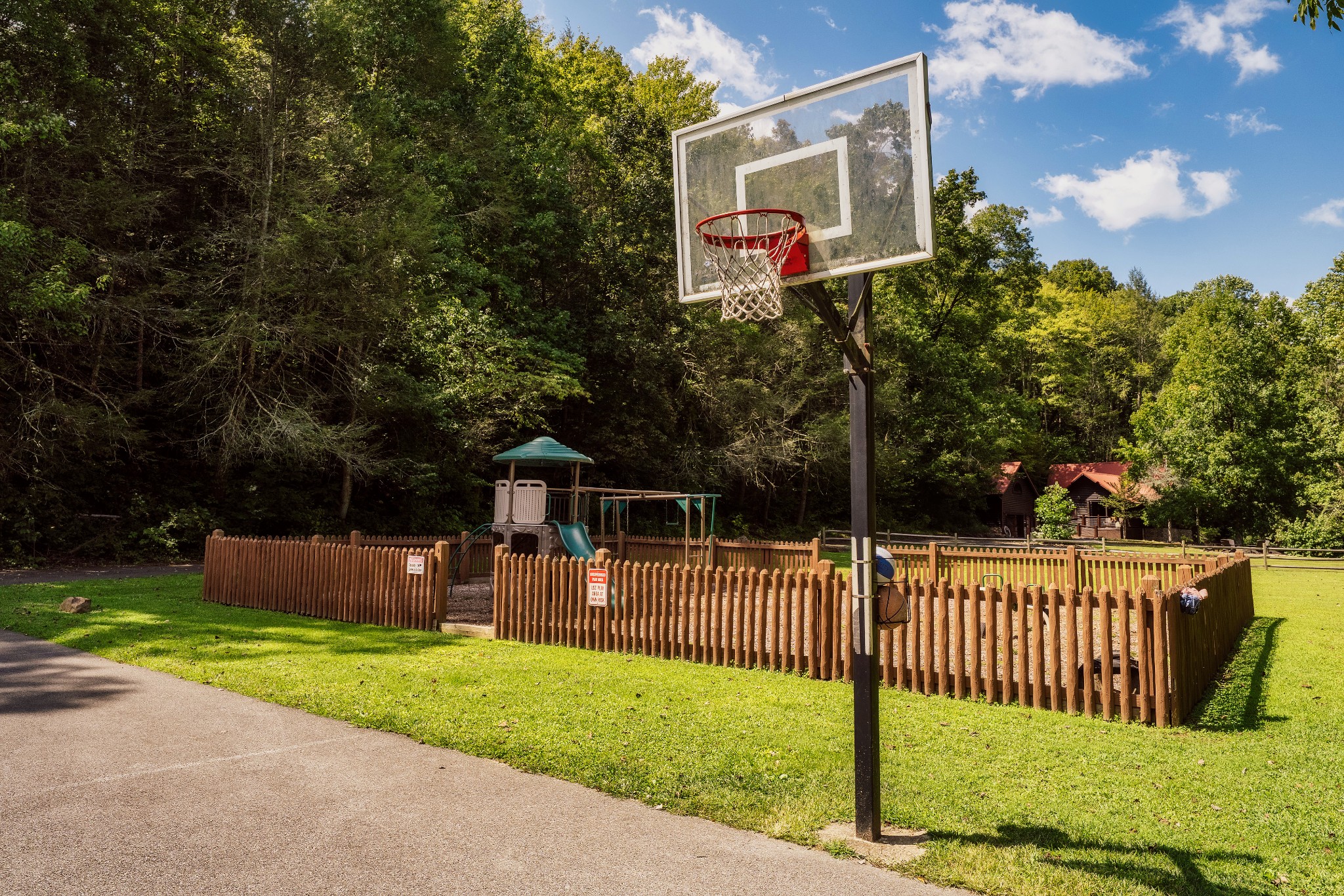 Basketball hoop and fenced playground beside wooded cabins.