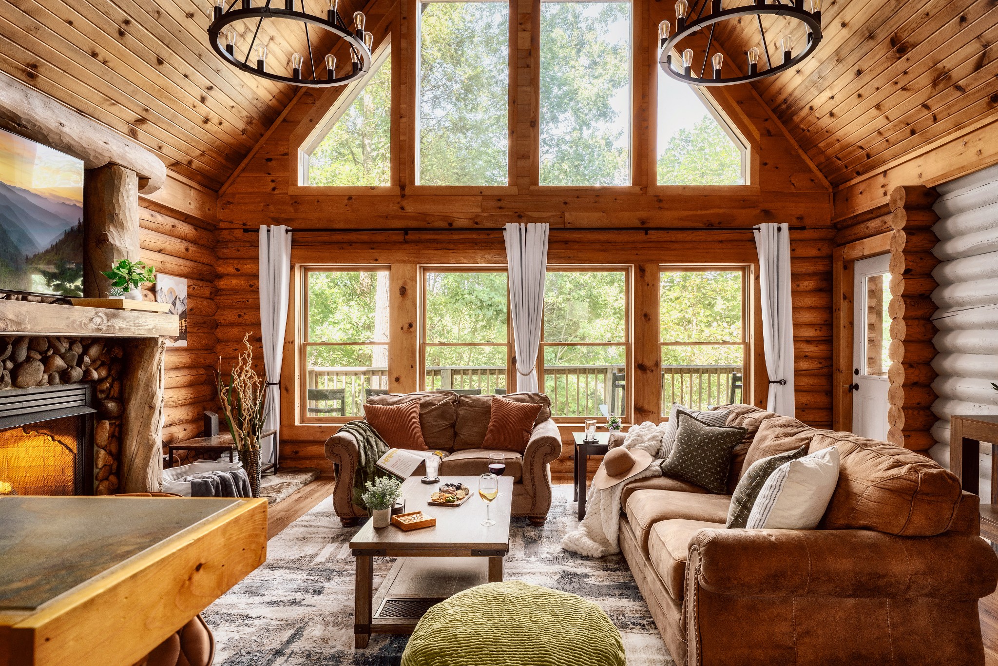 Log cabin living room with a stone fireplace, vaulted wooden ceiling, and large windows overlooking trees.