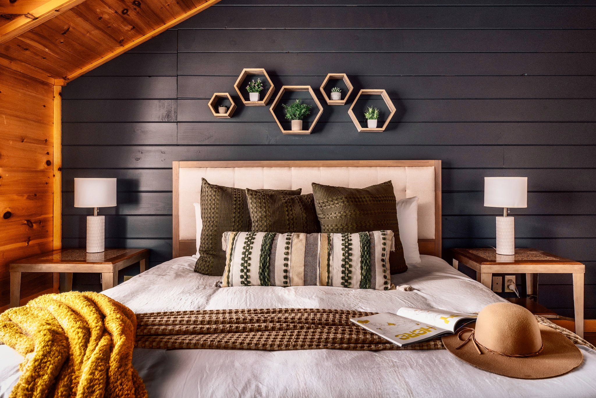 Furnished bedroom with a white bed, mustard throw, tan hat, and dark paneled accent wall.