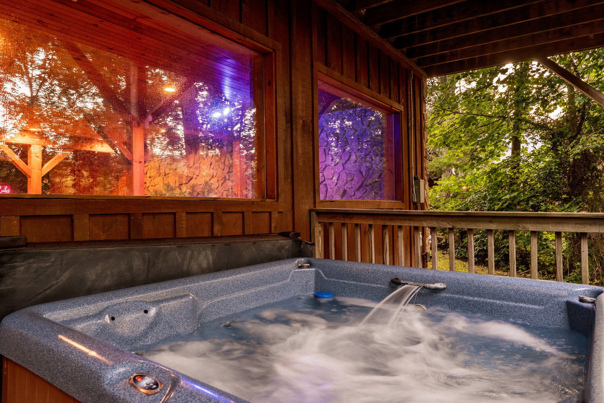 Outdoor hot tub on a covered wooden deck beside windows reflecting colorful lights.